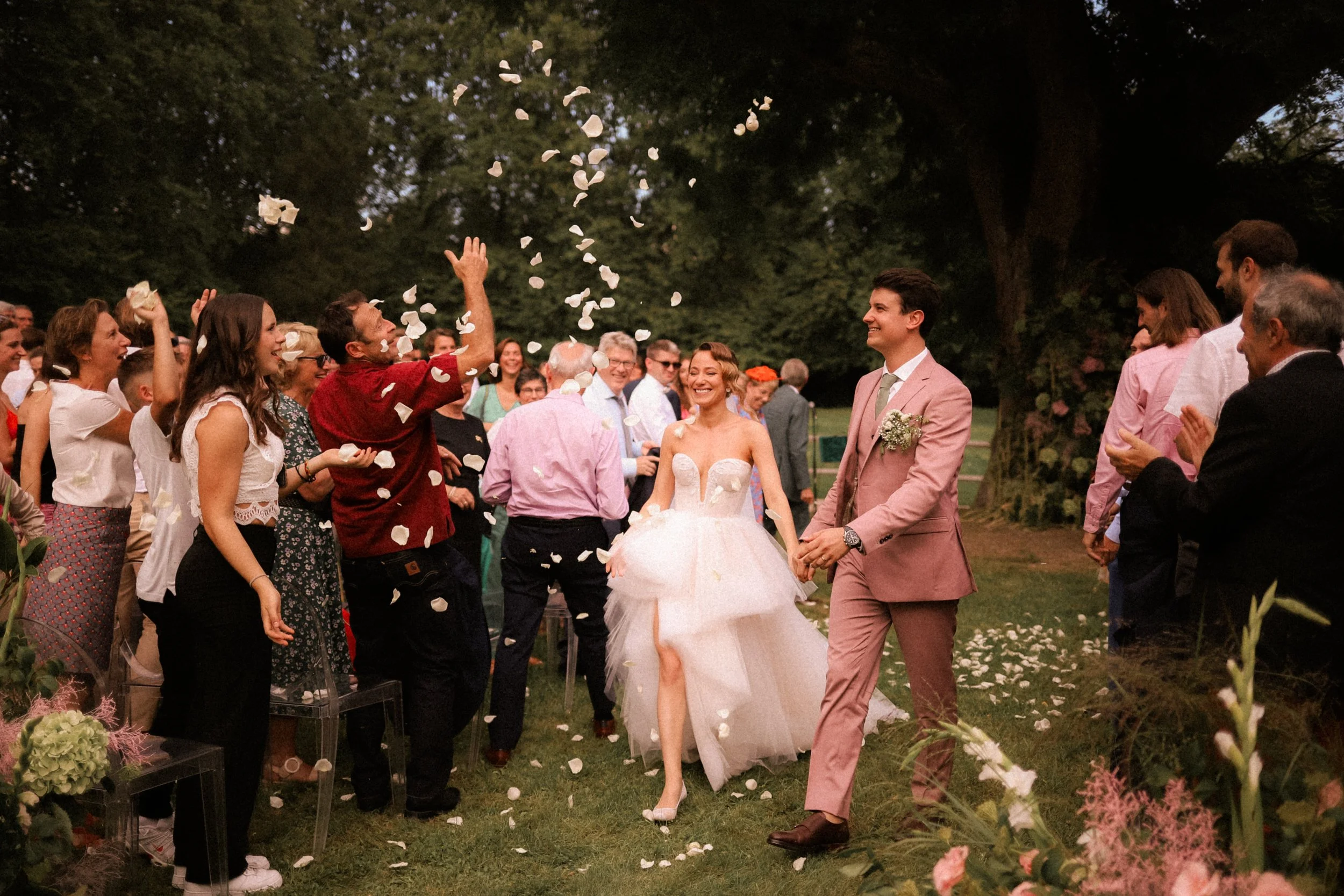 Un couple de mariés souriant à leur mariage en plein air, entourés d'invités lançant des pétales de fleurs, avec des arbres en arrière-plan et une ambiance joyeuse.