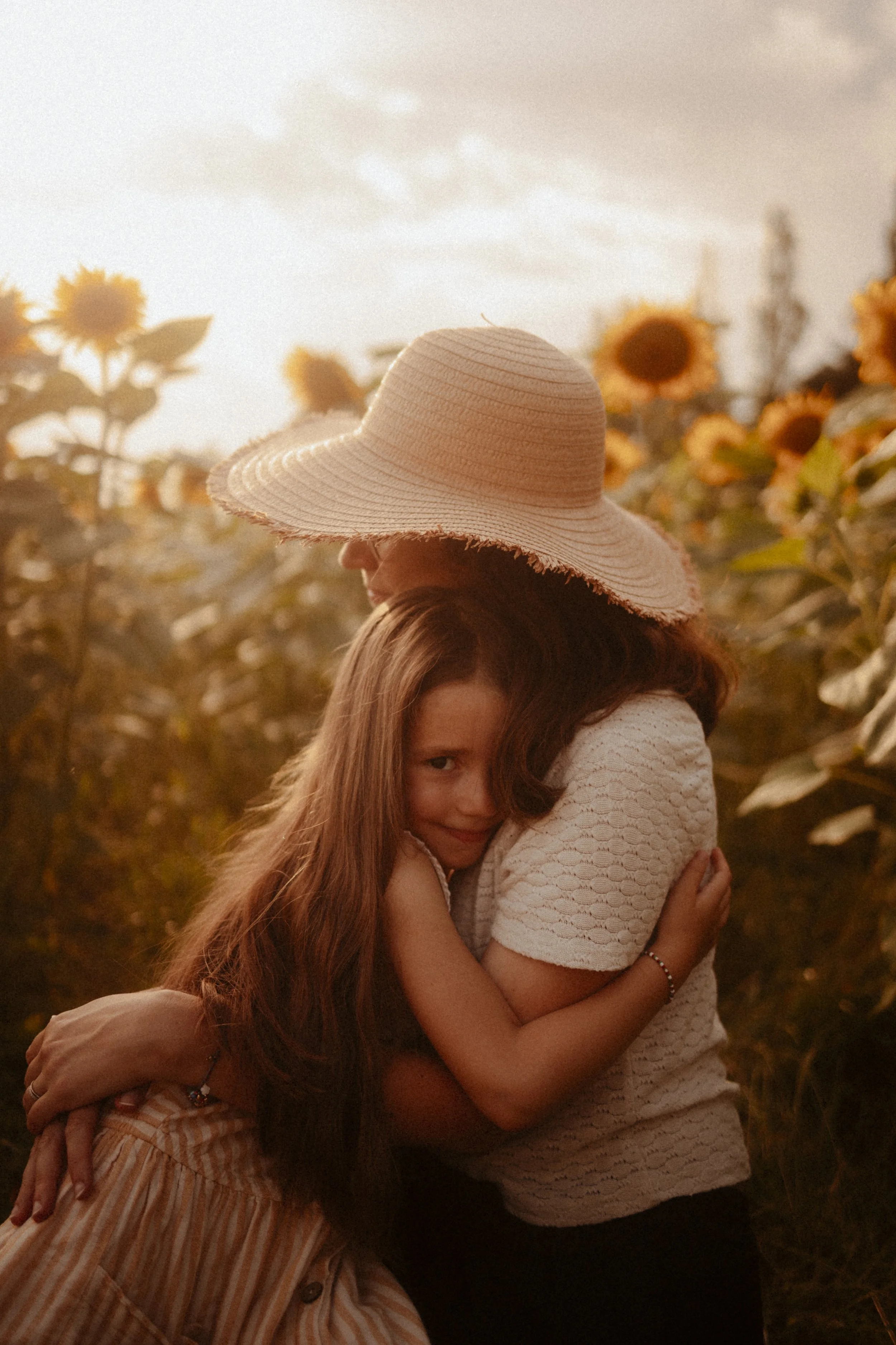 Une femme et une jeune fille s'embrassent dans un champ de tournesols au coucher du soleil, la femme porte un chapeau de paille large.