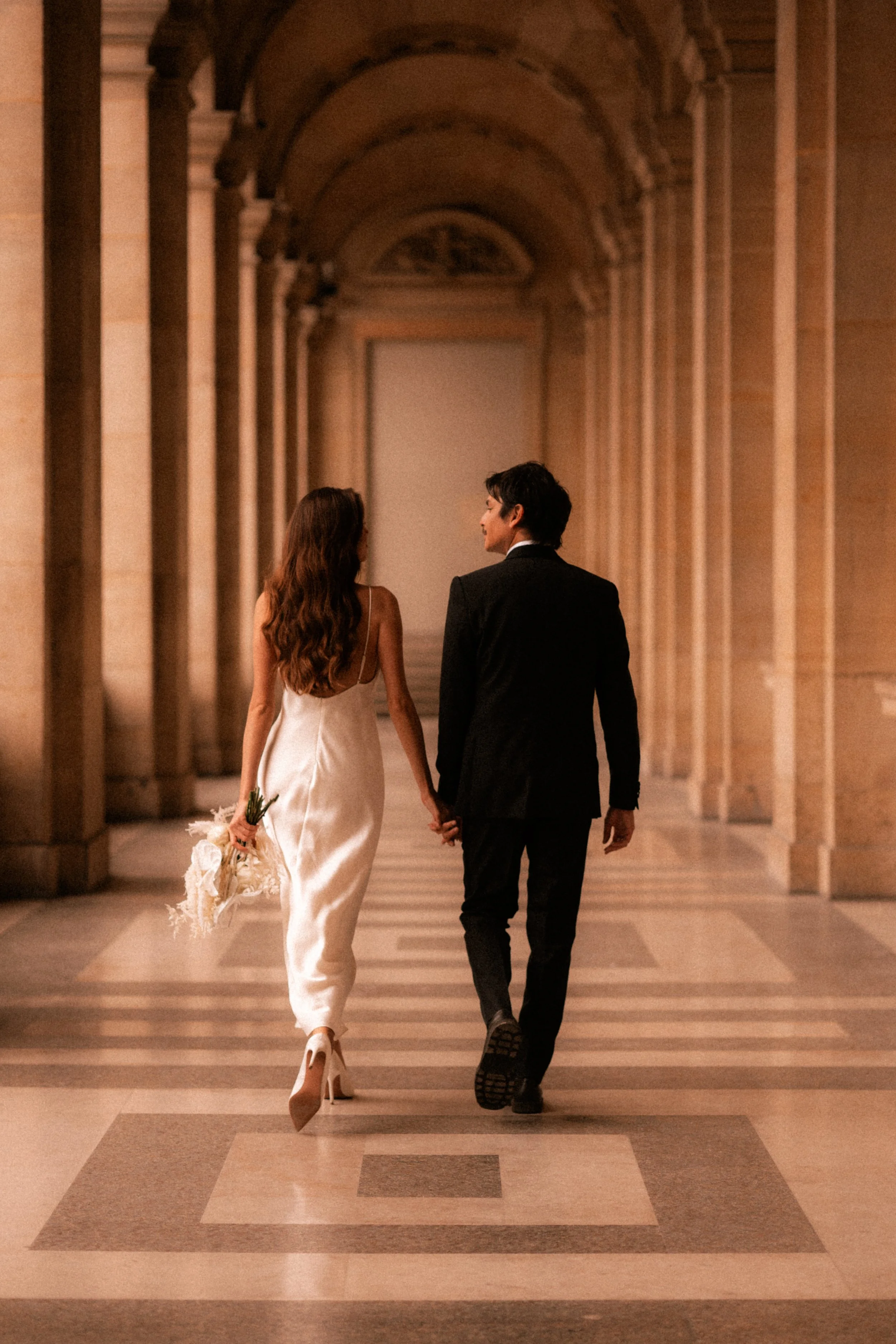 Un couple marchant main dans la main dans un couloir lumineux, l'homme porte un costume noir et la femme une robe blanche, tous deux vus de dos.