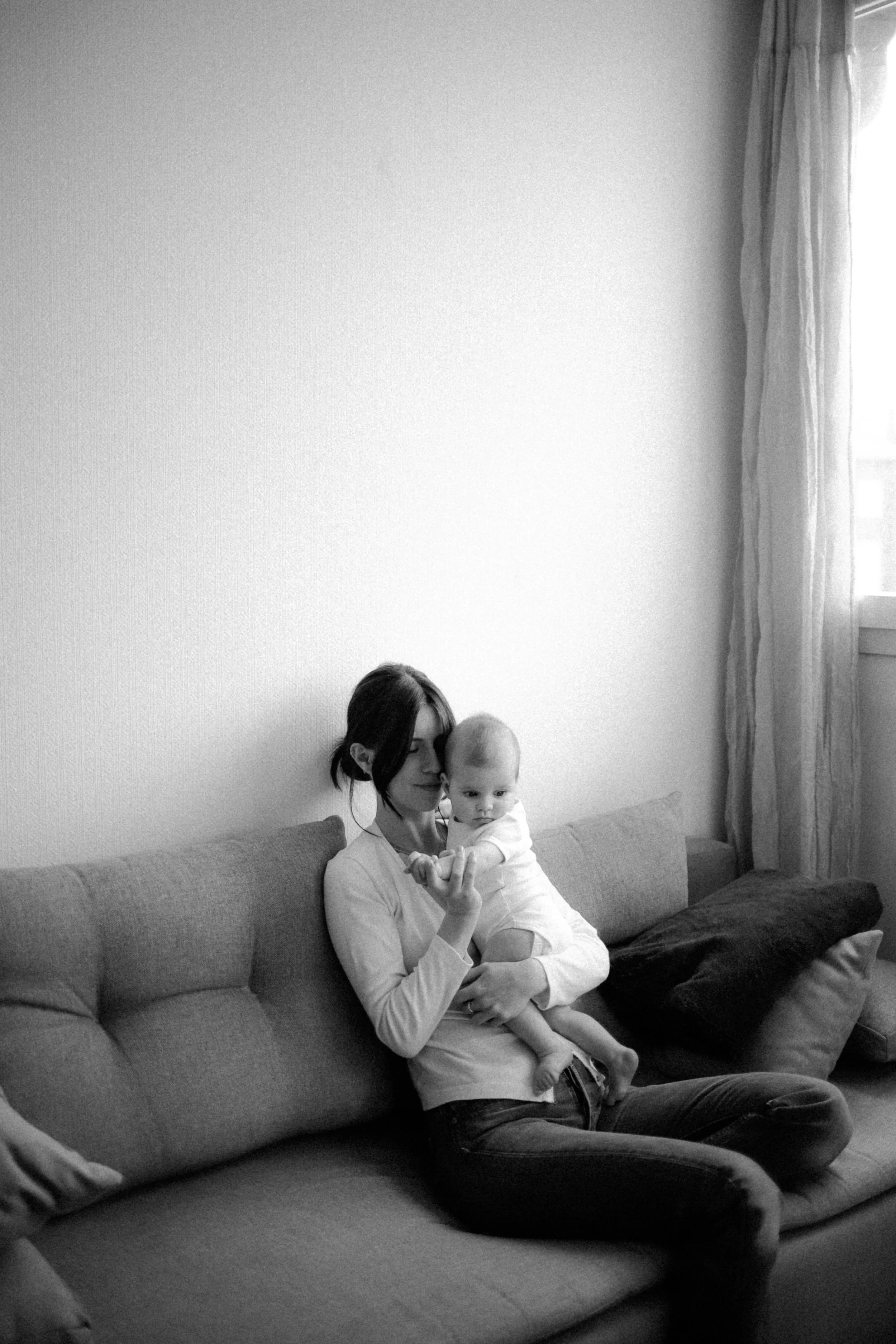 Une femme assise sur un canapé avec un enfant dans ses bras, dans une pièce lumineuse avec une fenêtre à droite.