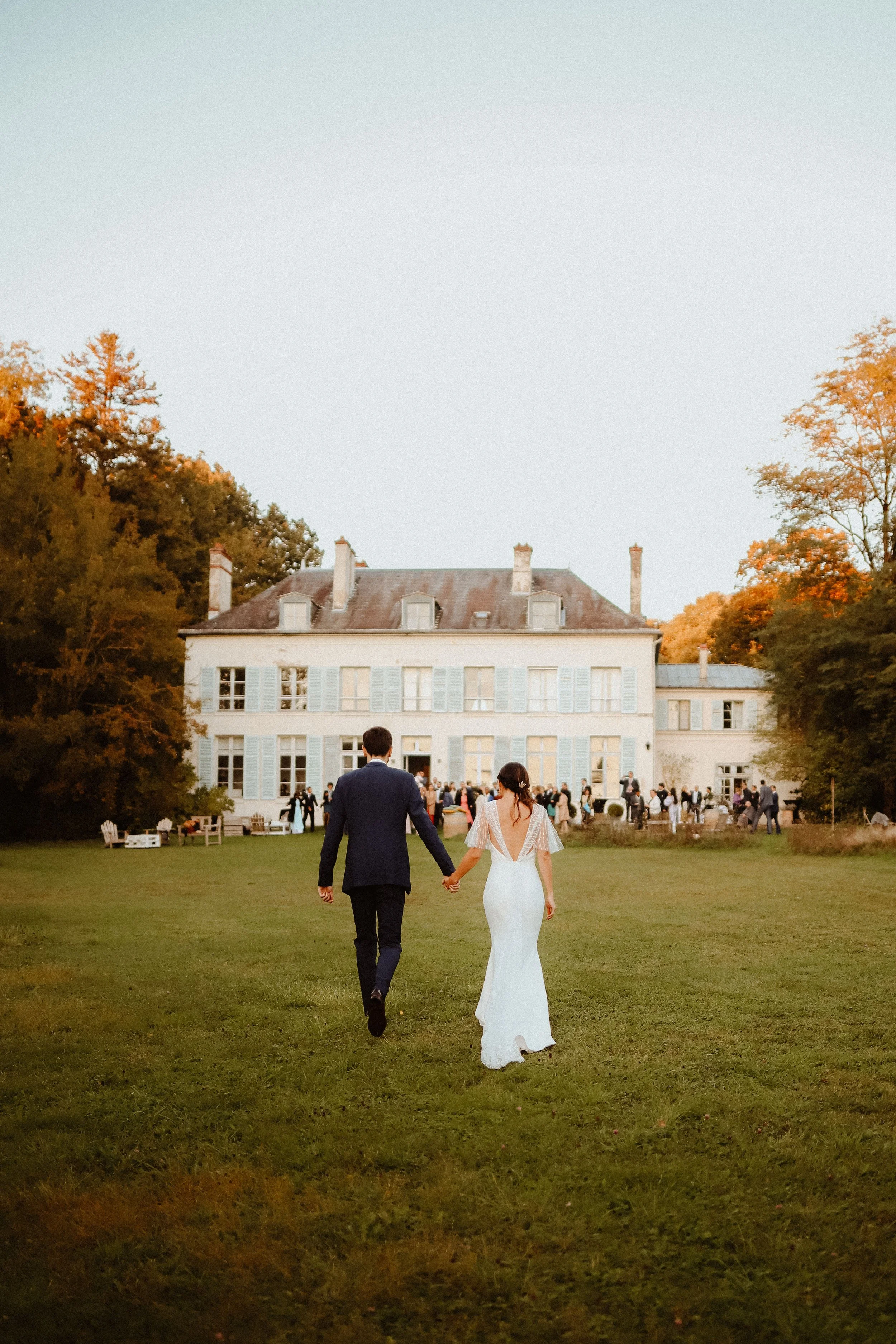 Un couple de mariés marchant main dans la main vers un château lors d'une cérémonie en plein air, entouré d'invités, avec des arbres aux couleurs d'automne.