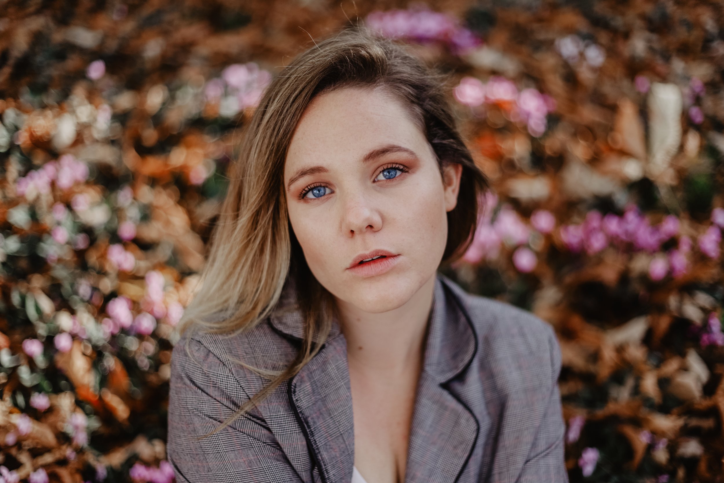 Jeune femme aux yeux bleus et cheveux longs, portant un blazer à carreaux, dans un environnement naturel avec des fleurs roses et des feuilles mortes au sol.