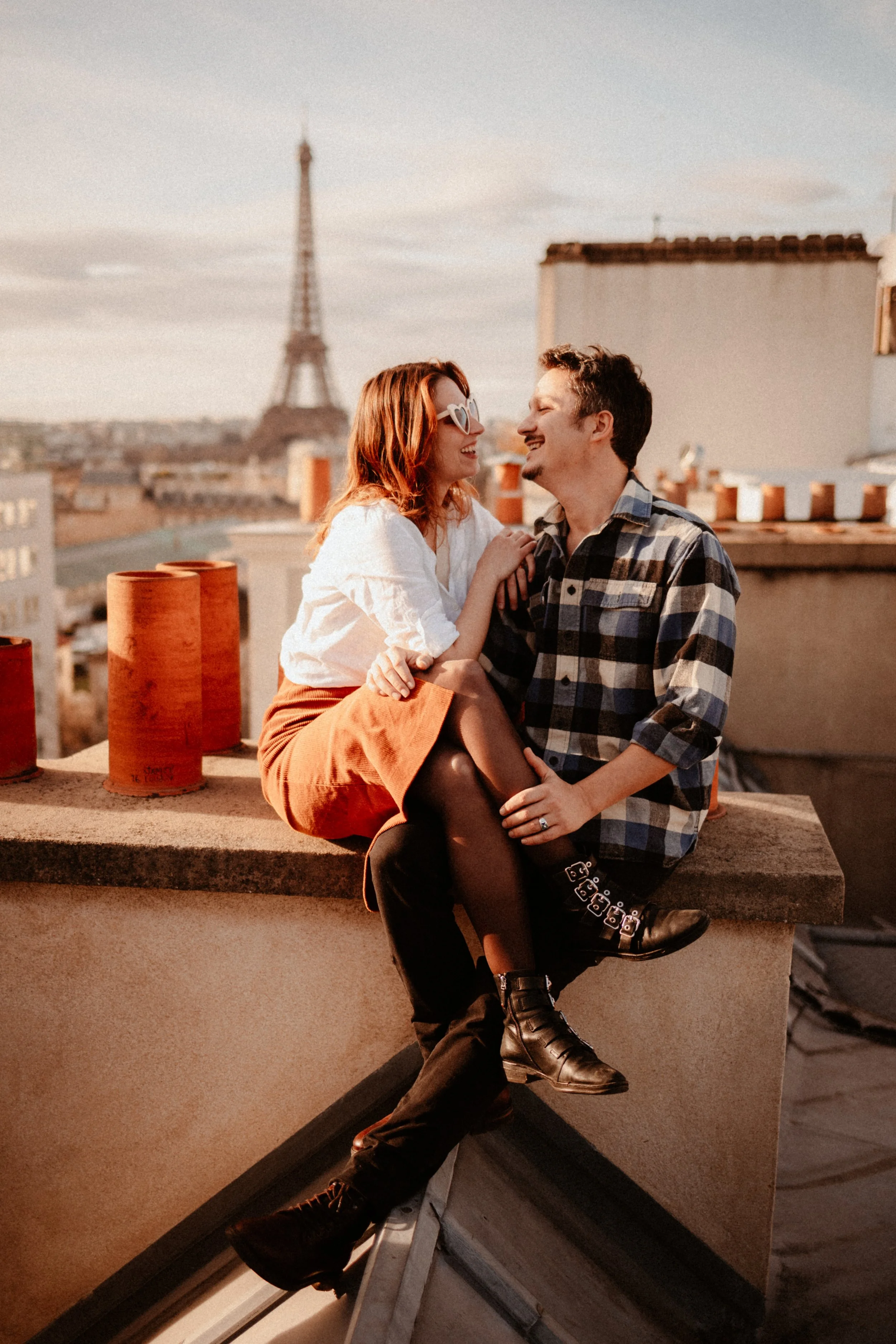 Un couple souriant assis sur un toit avec la Tour Eiffel en arrière-plan, pendant le coucher du soleil.