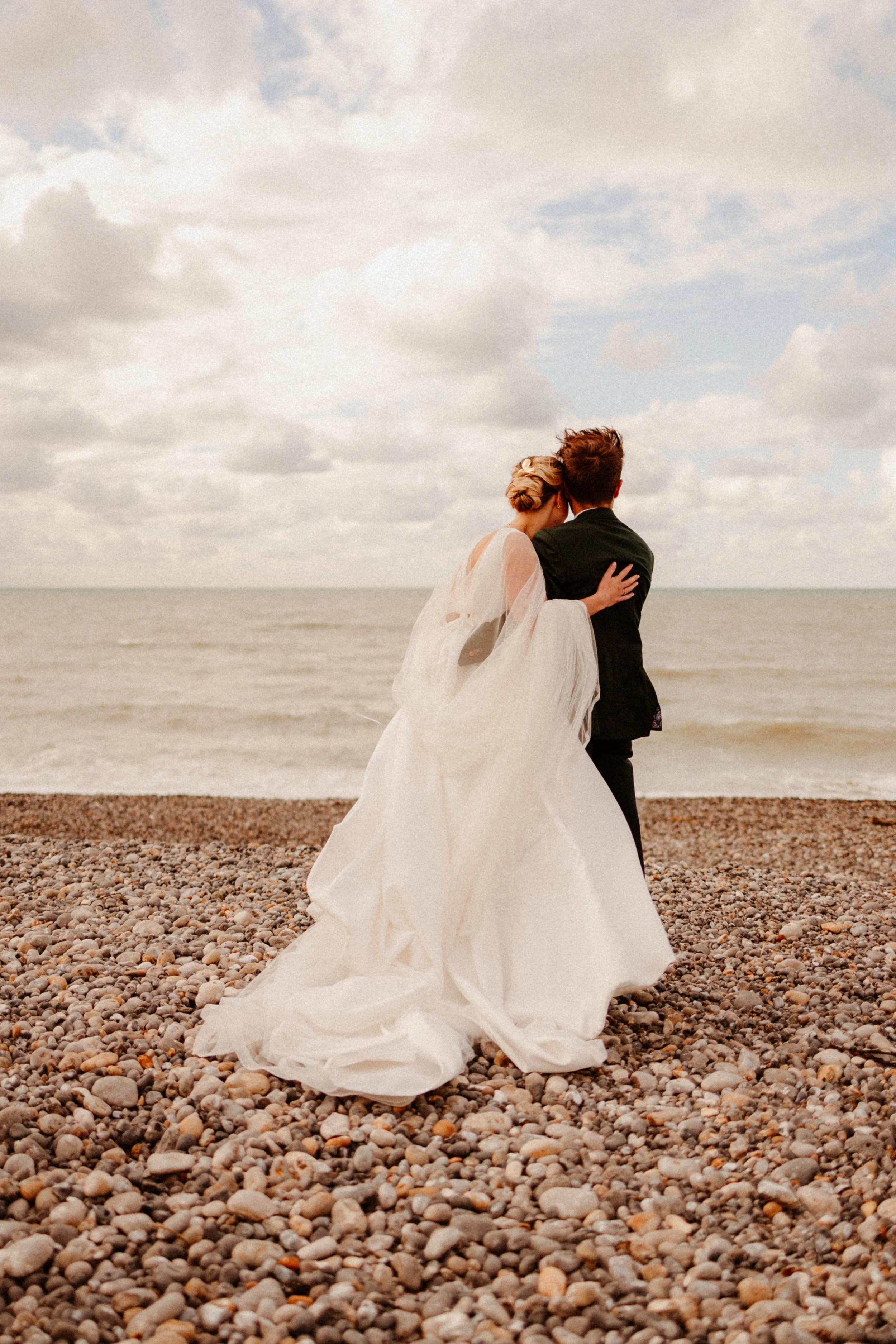 Un couple de mariés marche sur une plage de galets, avec la mer et un ciel nuageux en arrière-plan.