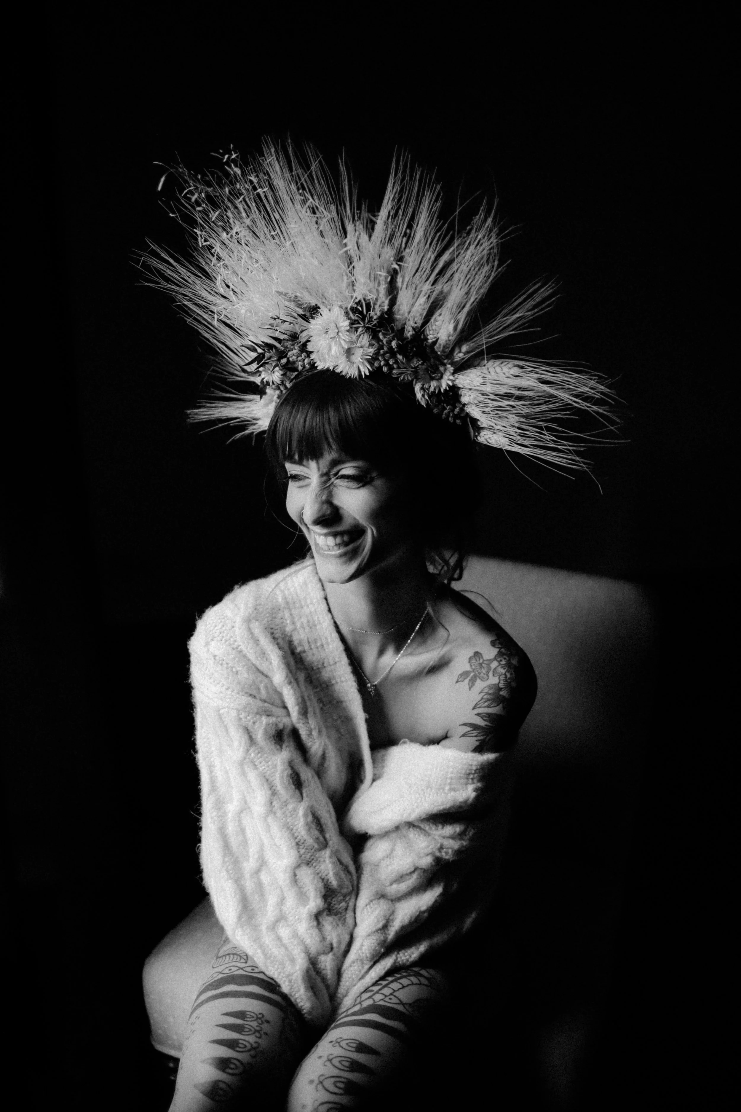 Une femme souriante portant une couronne de fleurs et de plumes, vêtue d'un pull en laine, avec des tatouages visibles sur l'épaule et les jambes, en noir et blanc.