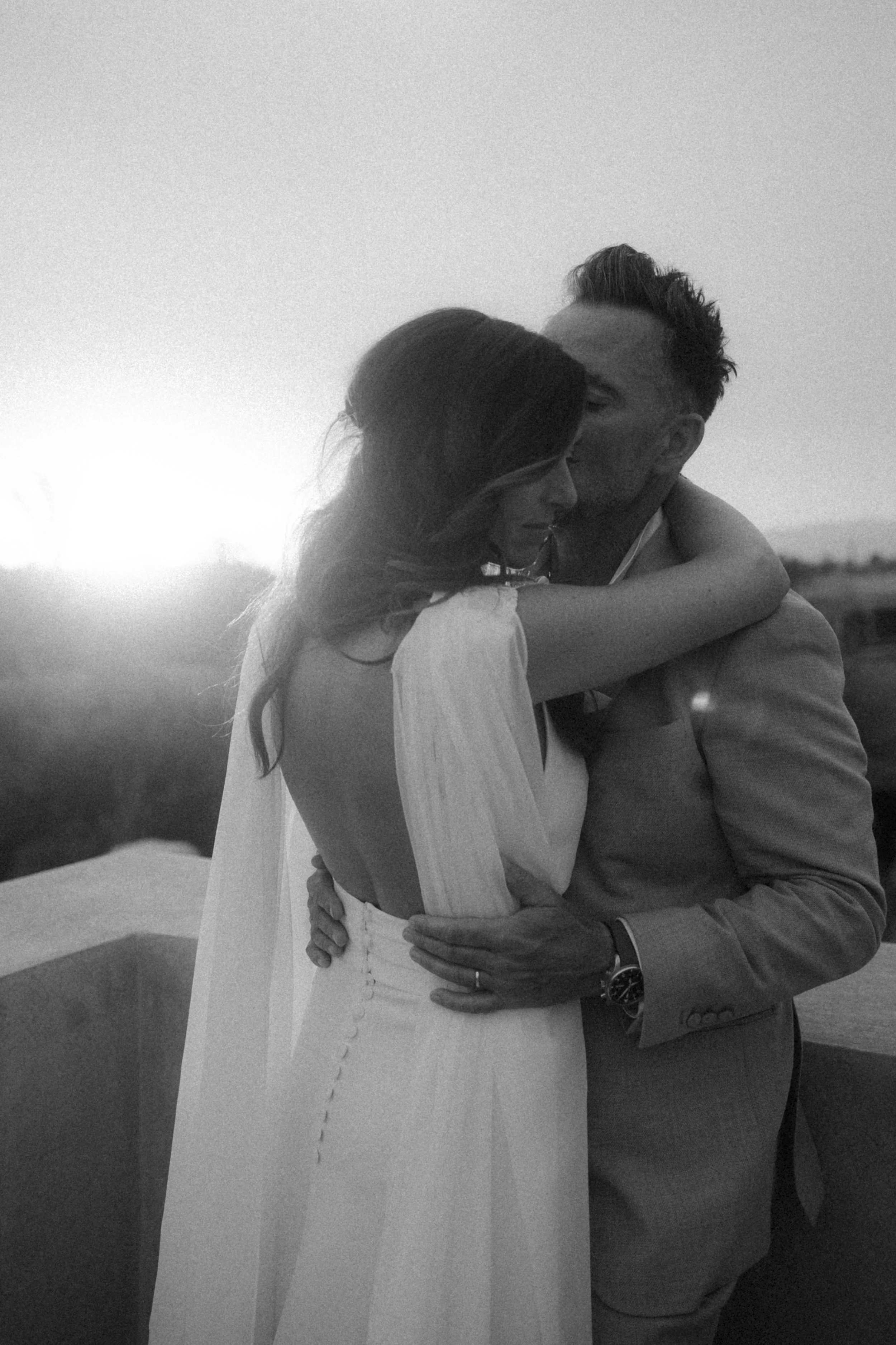 Un couple en tenue formelle s'embrassant lors d'un coucher de soleil en noir et blanc.