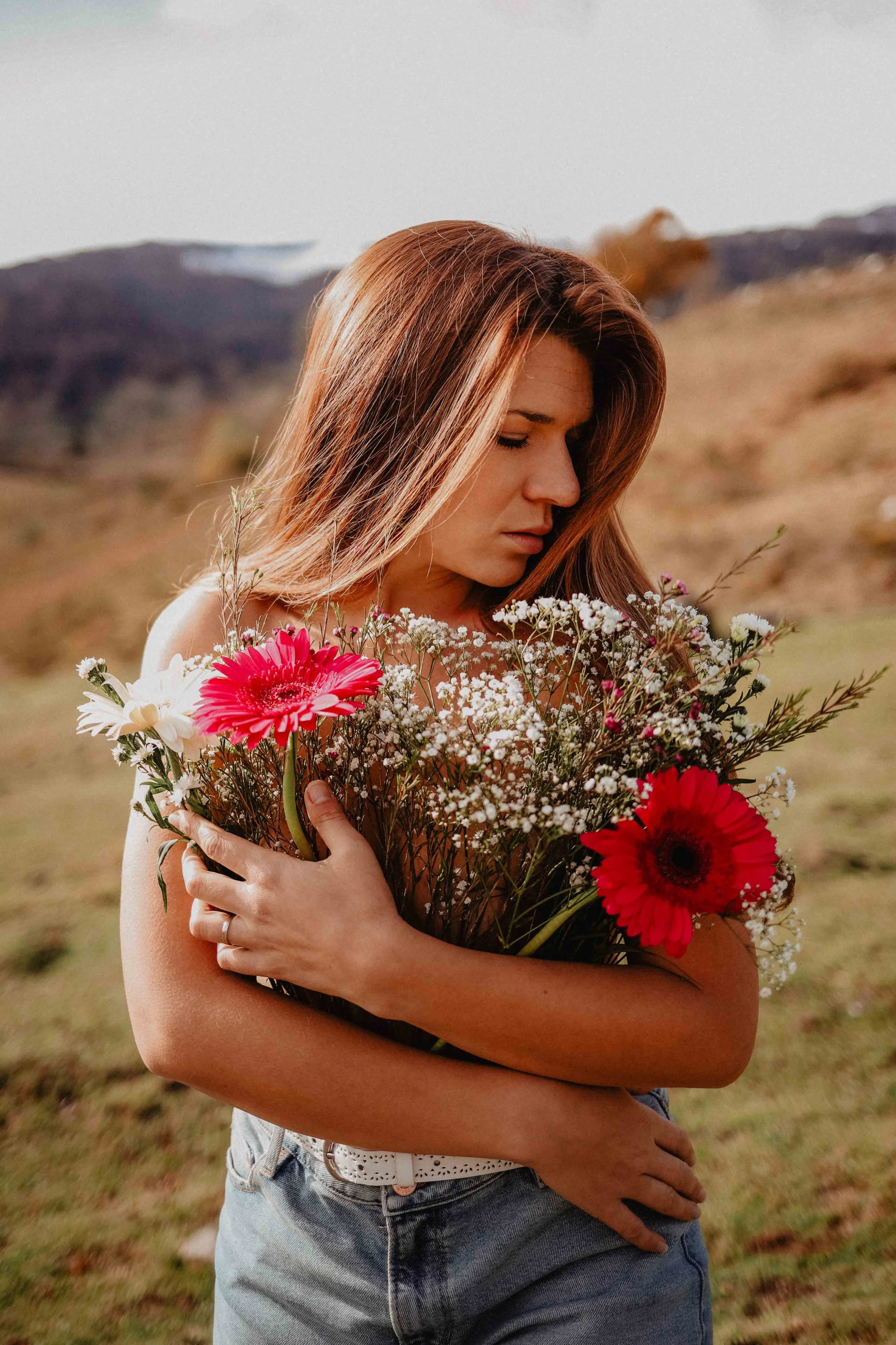 Jeune femme tenant un bouquet de fleurs, dont des gerberas rouges, dans un paysage champêtre.