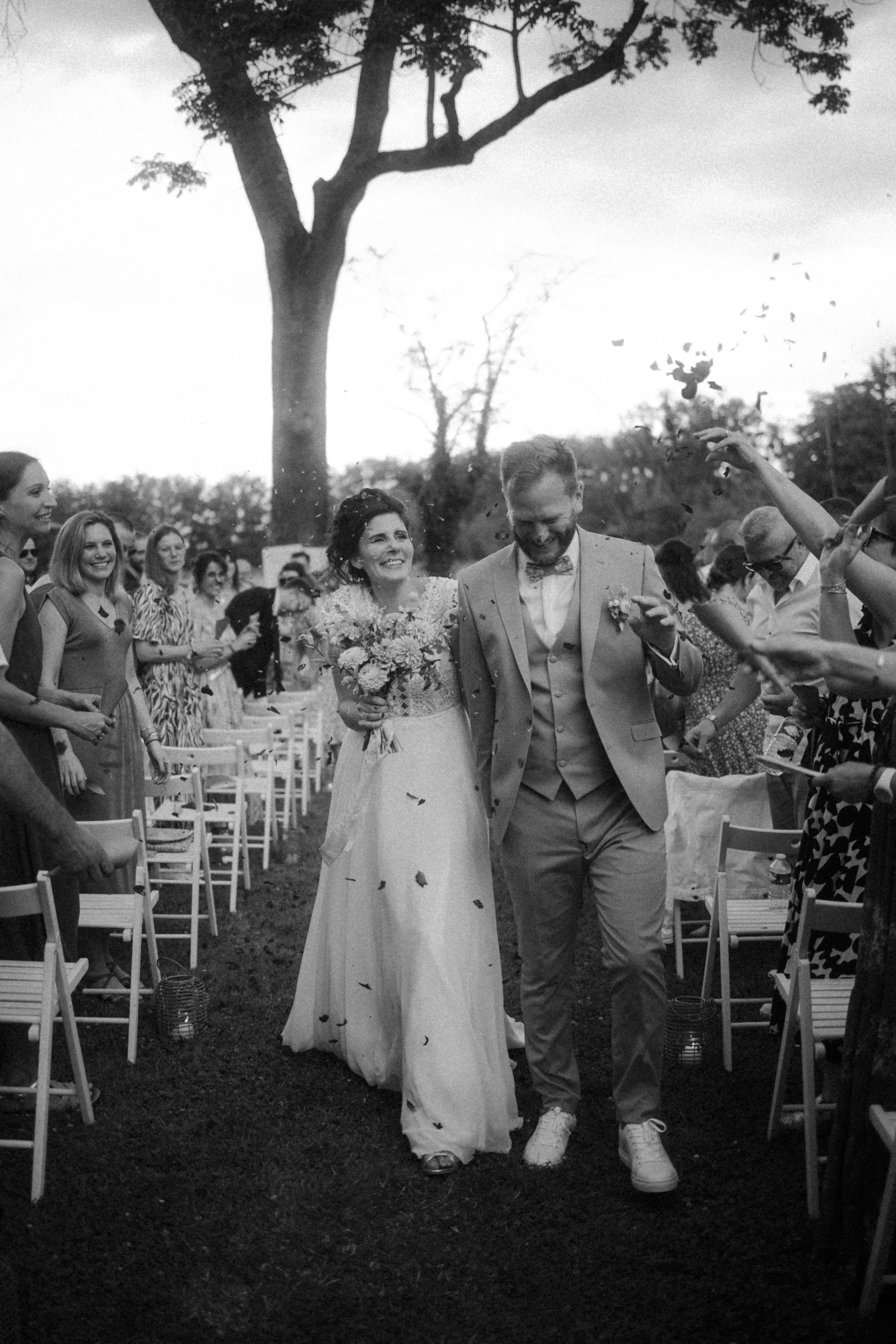 Un couple de mariés marche main dans la main lors de leur mariage en plein air, entourés d'invités qui célèbrent leur union, avec des arbres en arrière-plan.