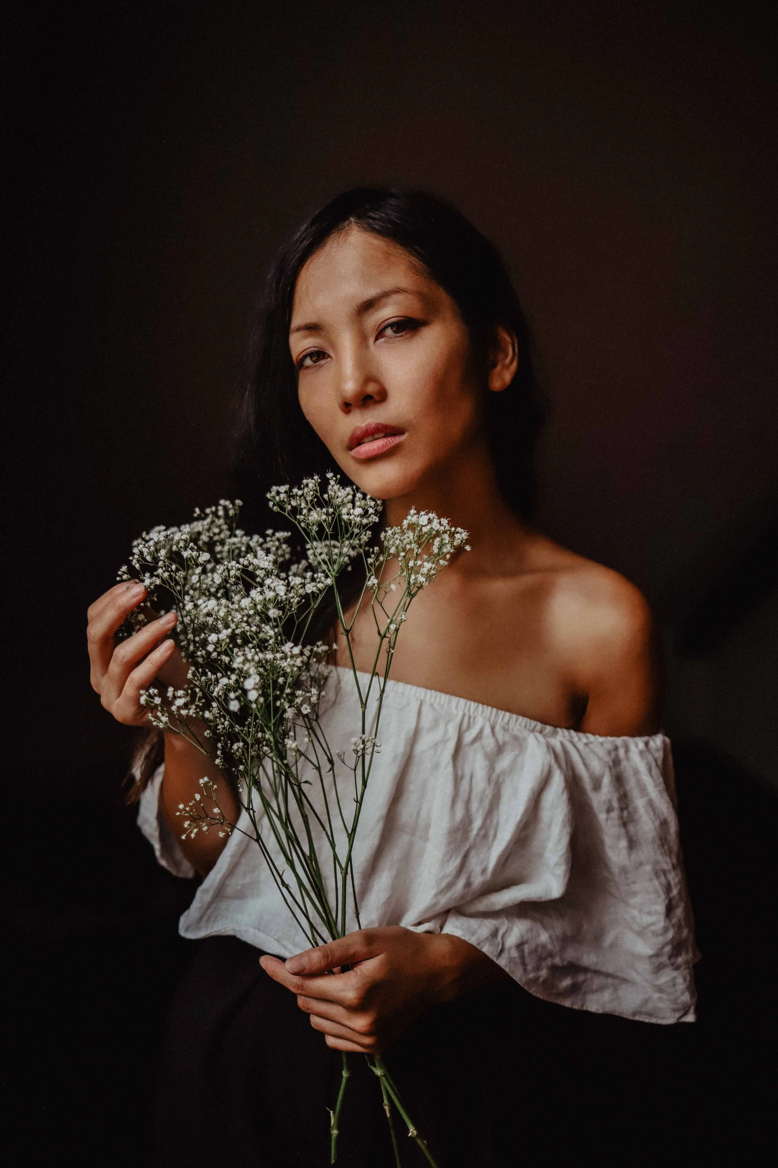 Une femme avec des cheveux noirs tenant un bouquet de petites fleurs blanches contre un fond sombre.