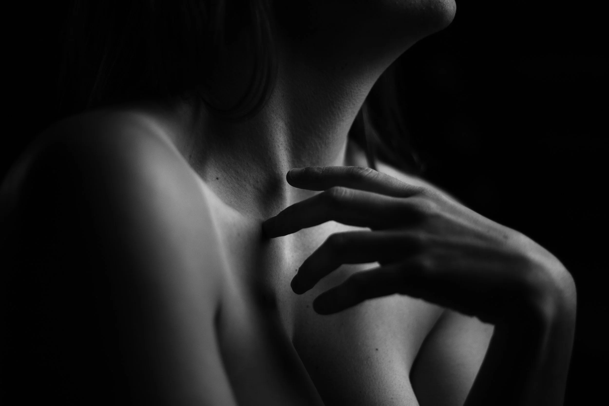 Photographie en noir et blanc d'une femme dont la main touche la clavicule, avec une lumière douce mettant en valeur la texture de la peau.