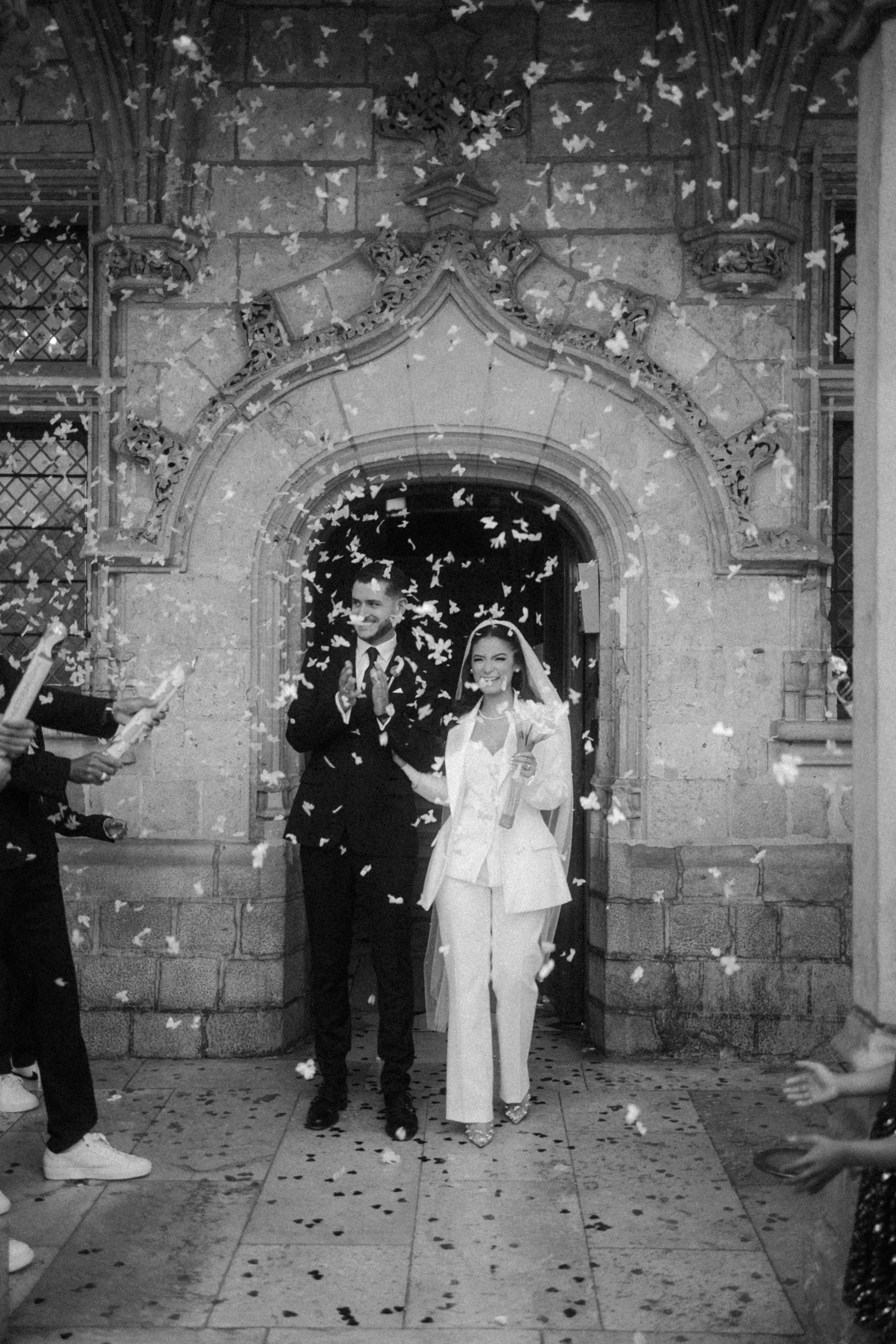 Un couple de mariés souriants sortant de l'église sous une pluie de confettis, entourés de personnes qui lancent des confettis, dans une scène en noir et blanc.