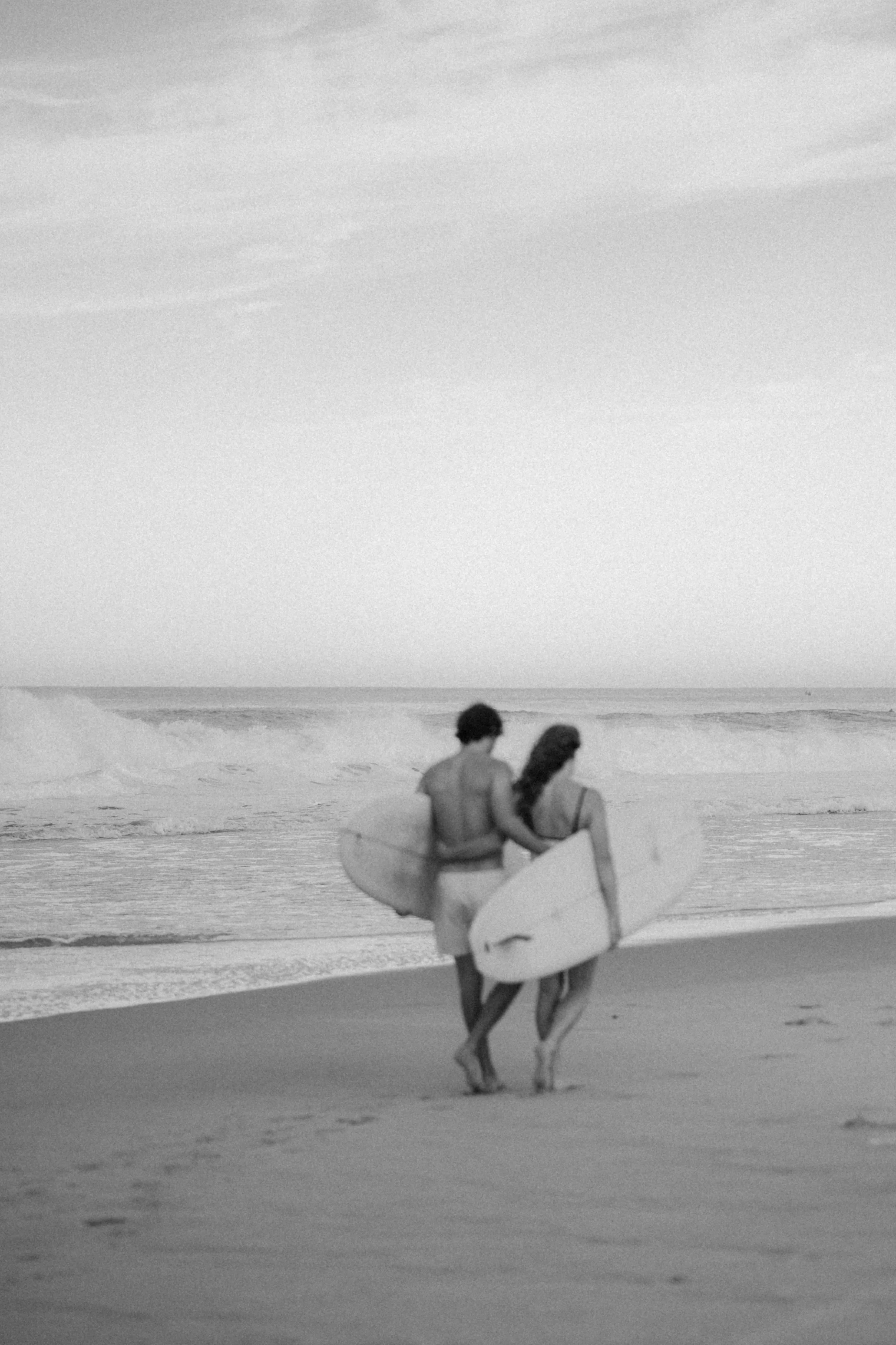 Deux surfeurs marche sur la plage en direction de l'océan avec leurs planches de surf, photographiés en noir et blanc.