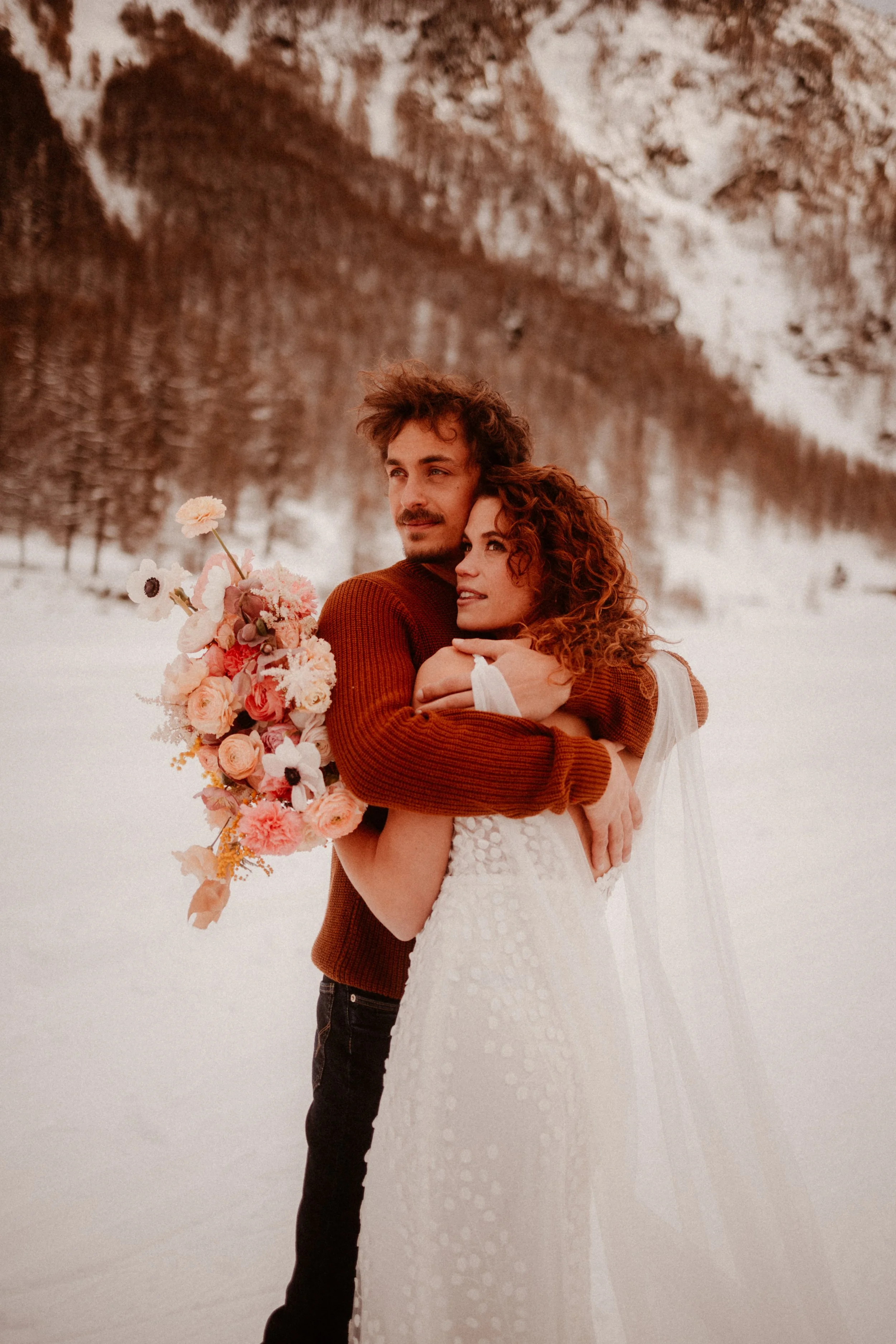Un couple en habits de mariage dans un paysage enneigé, la femme tient un bouquet de fleurs et le homme l'enlace.