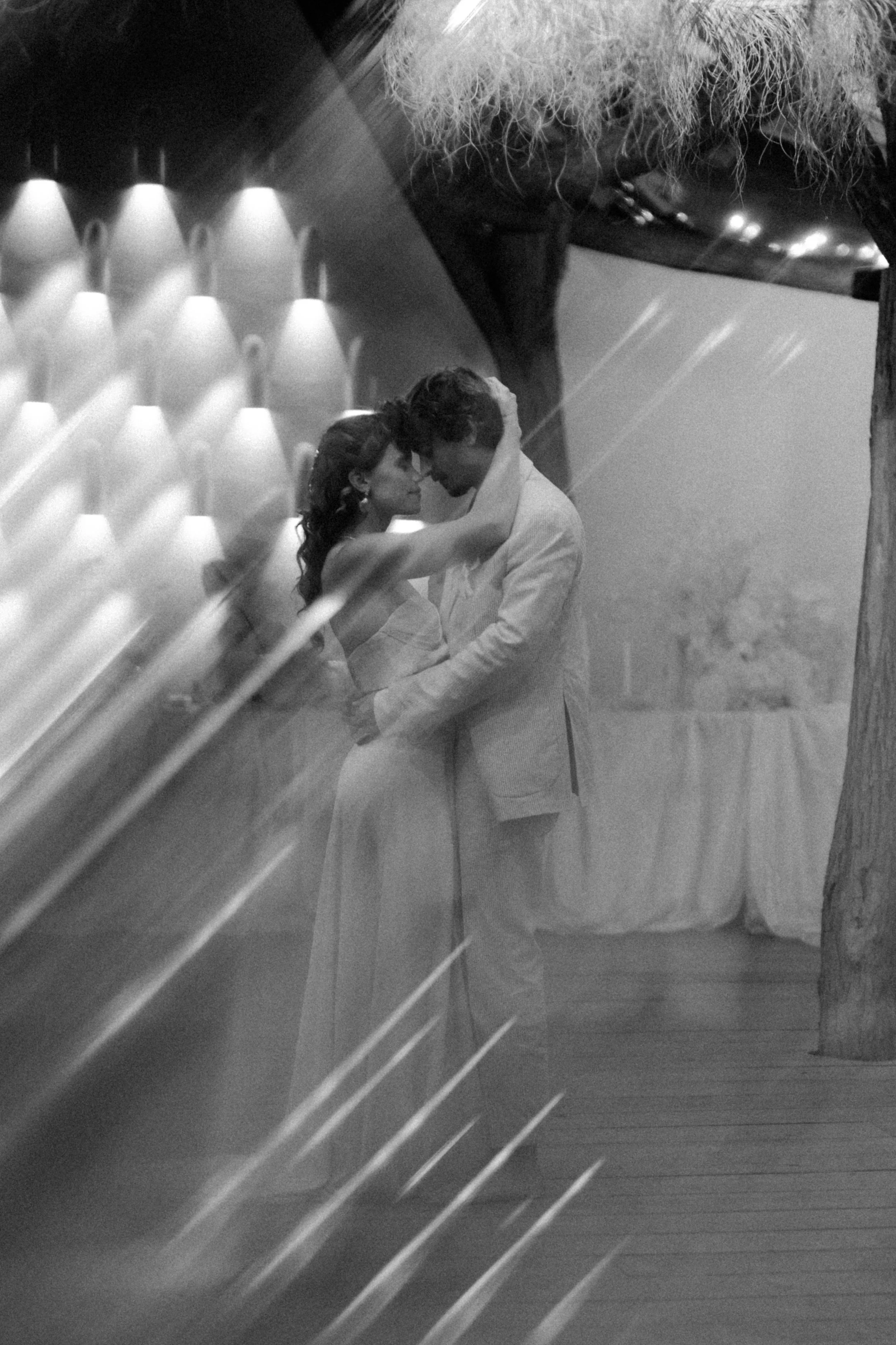 Un couple en tenue de mariage danse dans une ambiance romantique, entouré de lumières tamisées et décorations. La photo est en noir et blanc.