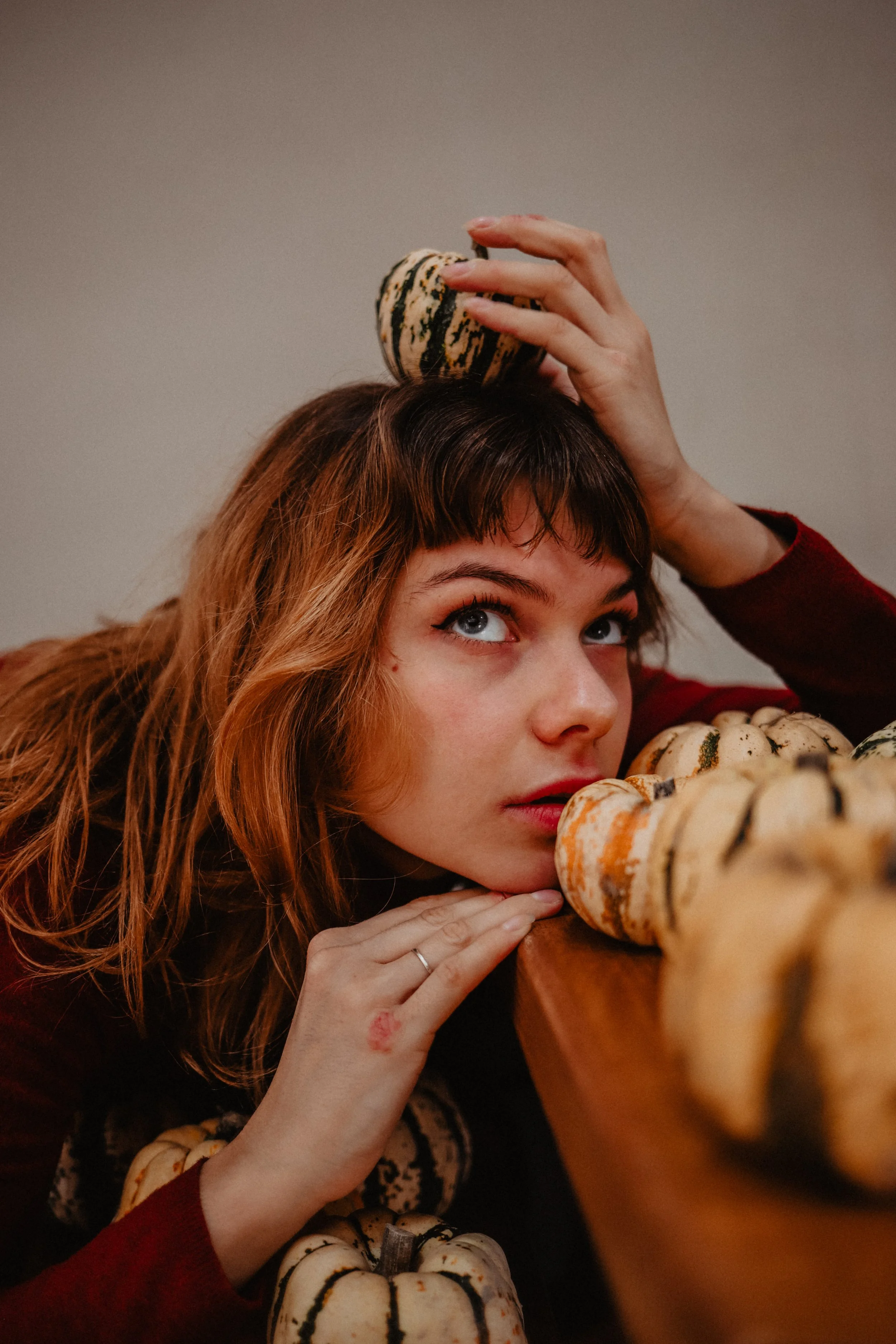 Jeune femme aux cheveux roux, reposant sa tête et son menton sur une surface en bois, entourée de petits potirons de différentes couleurs et motifs, avec un potiron posé au sommet de sa tête.