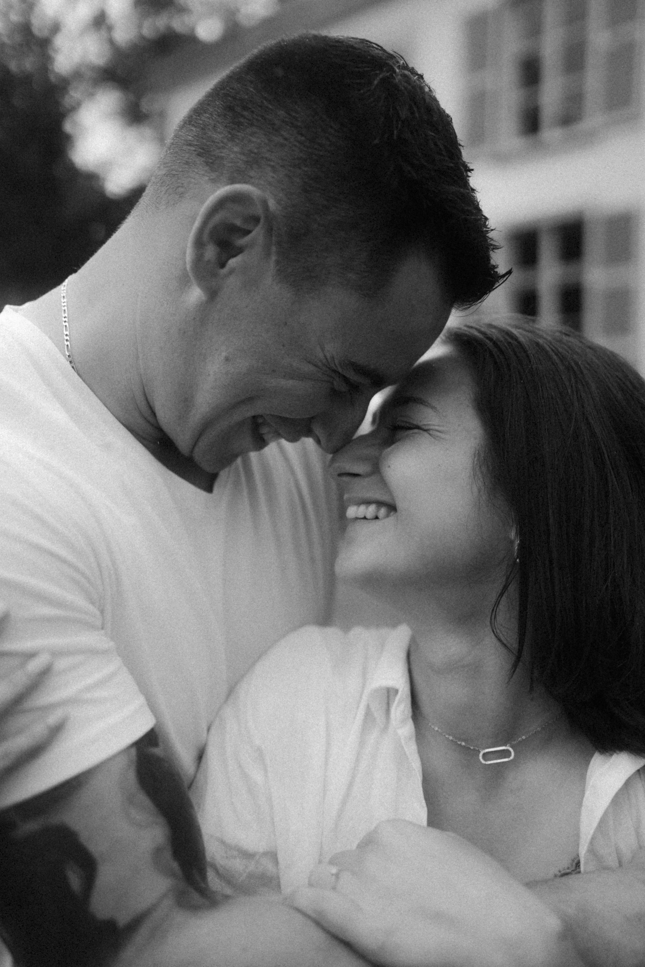 Un couple souriant en se tenant la tête en se touchant le front, dans un cadre extérieur, en noir et blanc.
