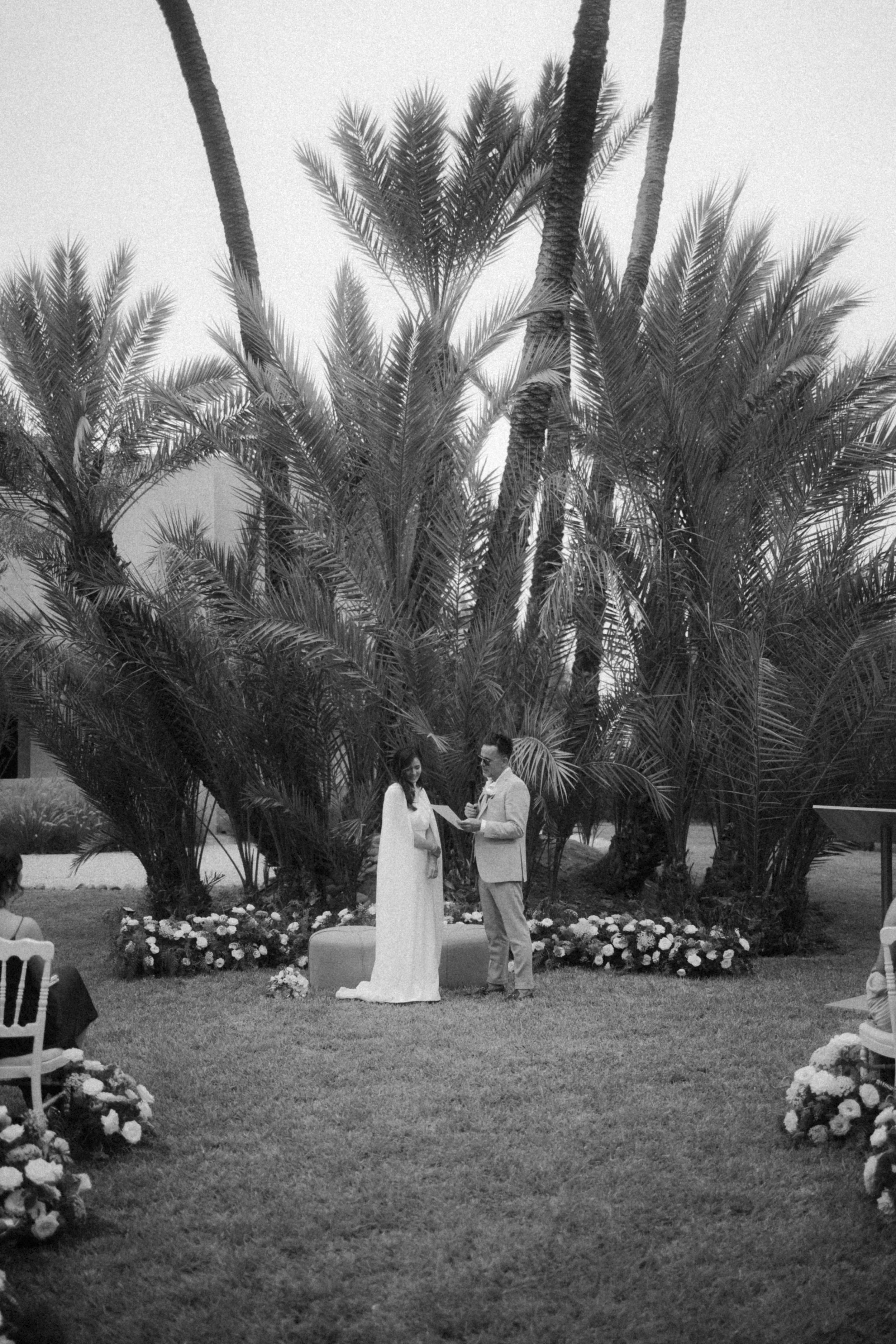 Cérémonie de mariage en plein air avec couple échangeant leurs vœux sous de grands palmiers, entourés de fleurs et d'une audience assise.