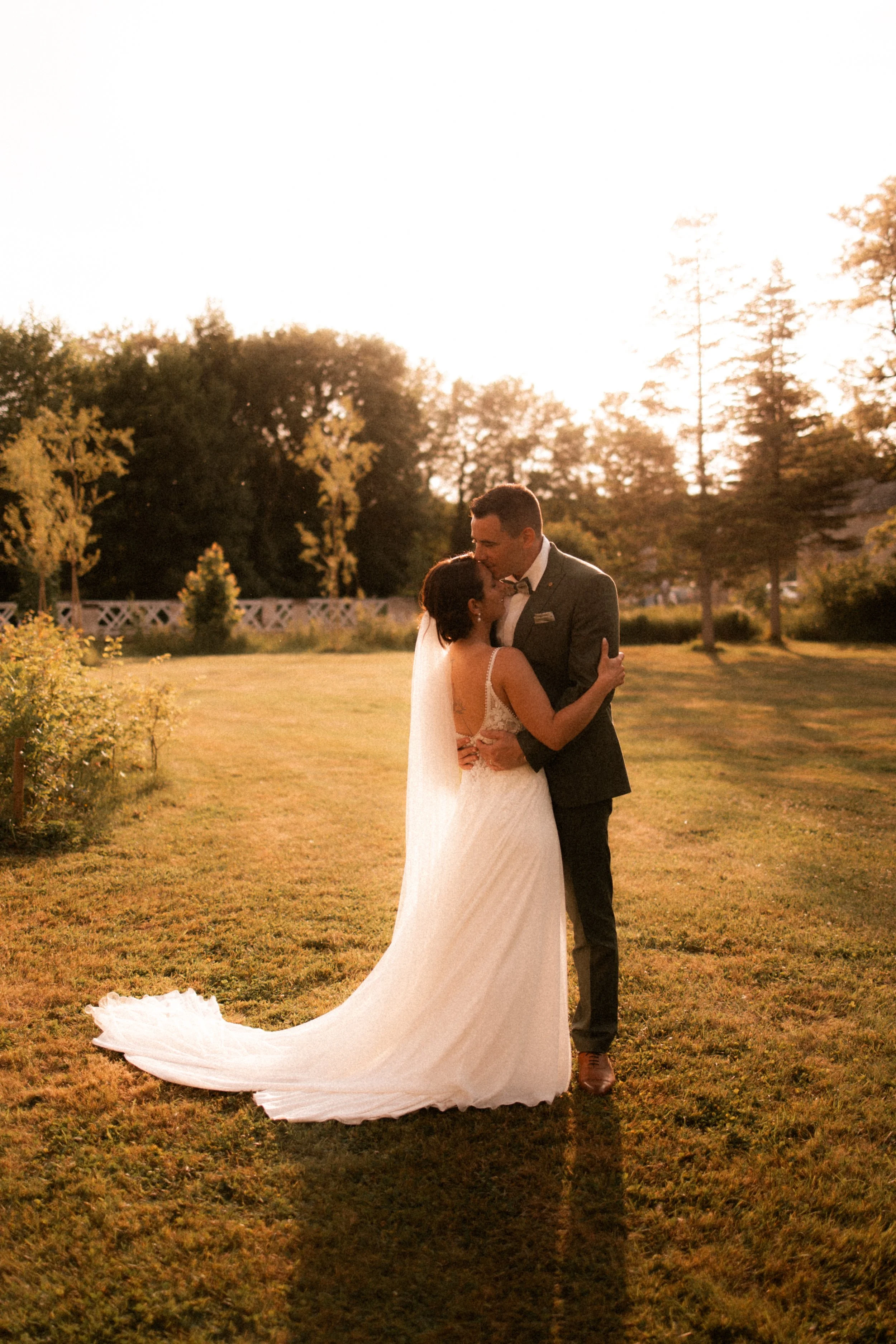 Un couple de mariés s'embrassant dans un jardin en soirée, portant une robe de mariée blanche et un costume sombre, avec le soleil couchant en arrière-plan.