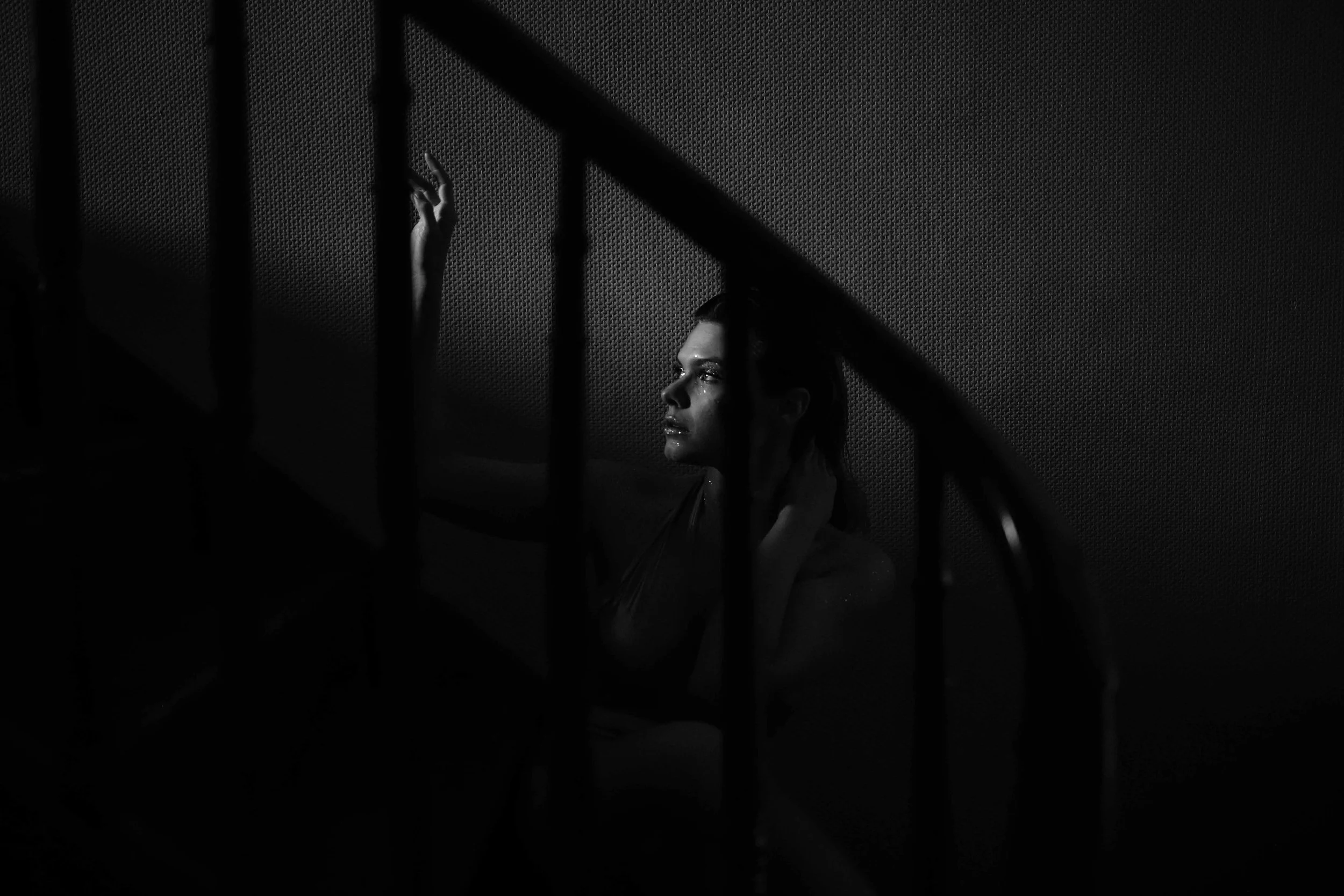 Une femme assise dans un environnement sombre, éclairée partiellement, vue à travers des barreaux d'escalier.