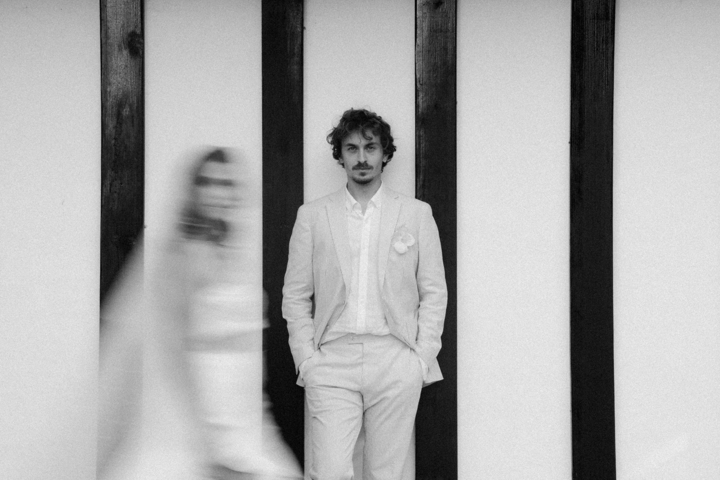 Un homme debout, vêtu d'un costume clair, pose contre un mur en bois clair et foncé, dans une image en noir et blanc. Une femme passe devant lui, floue, dans le premier plan.
