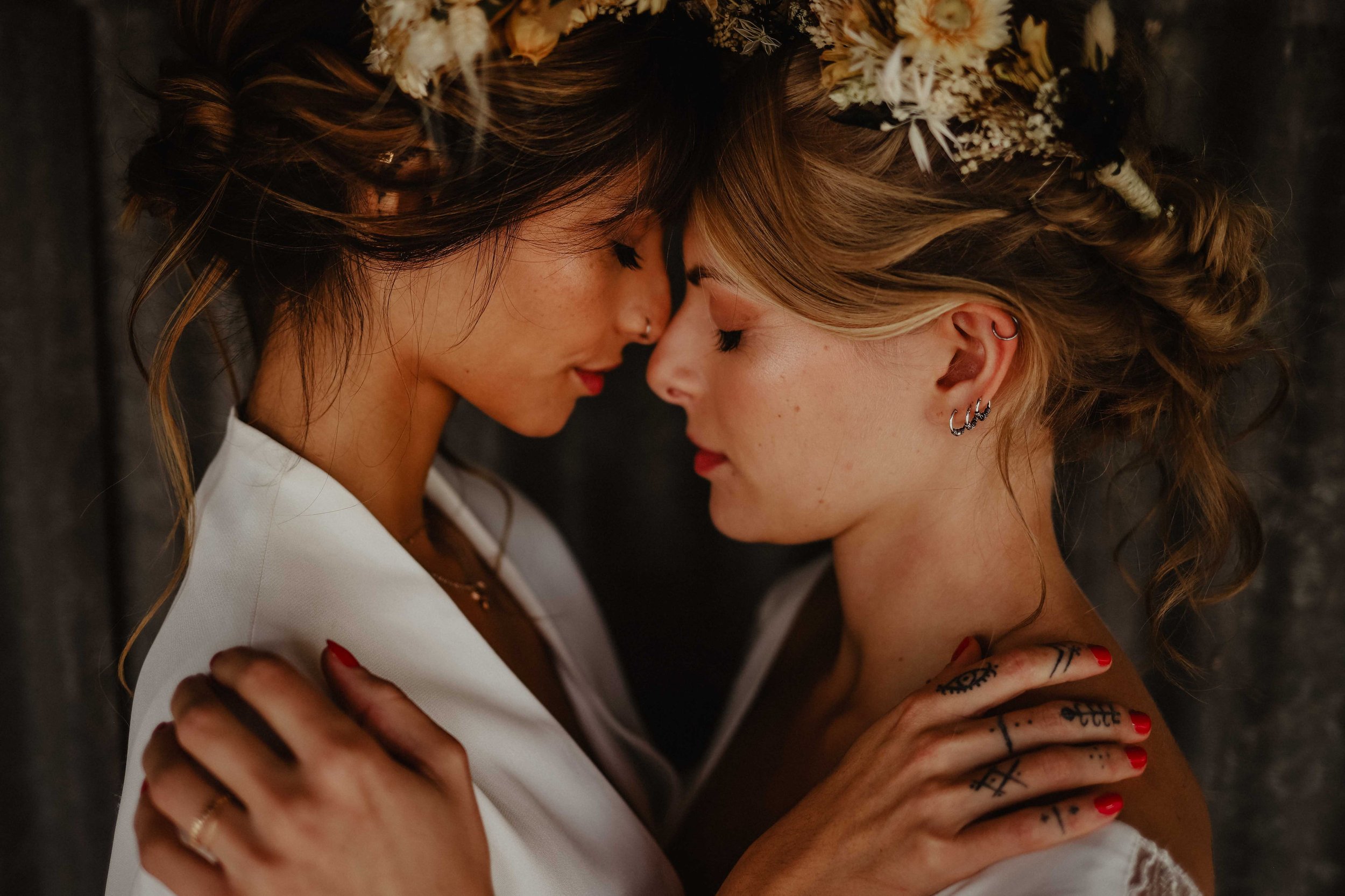 Deux femmes avec des couronnes de fleurs se tiennent proches, les fronts touchés, dans une pose intime, sur fond sombre.
