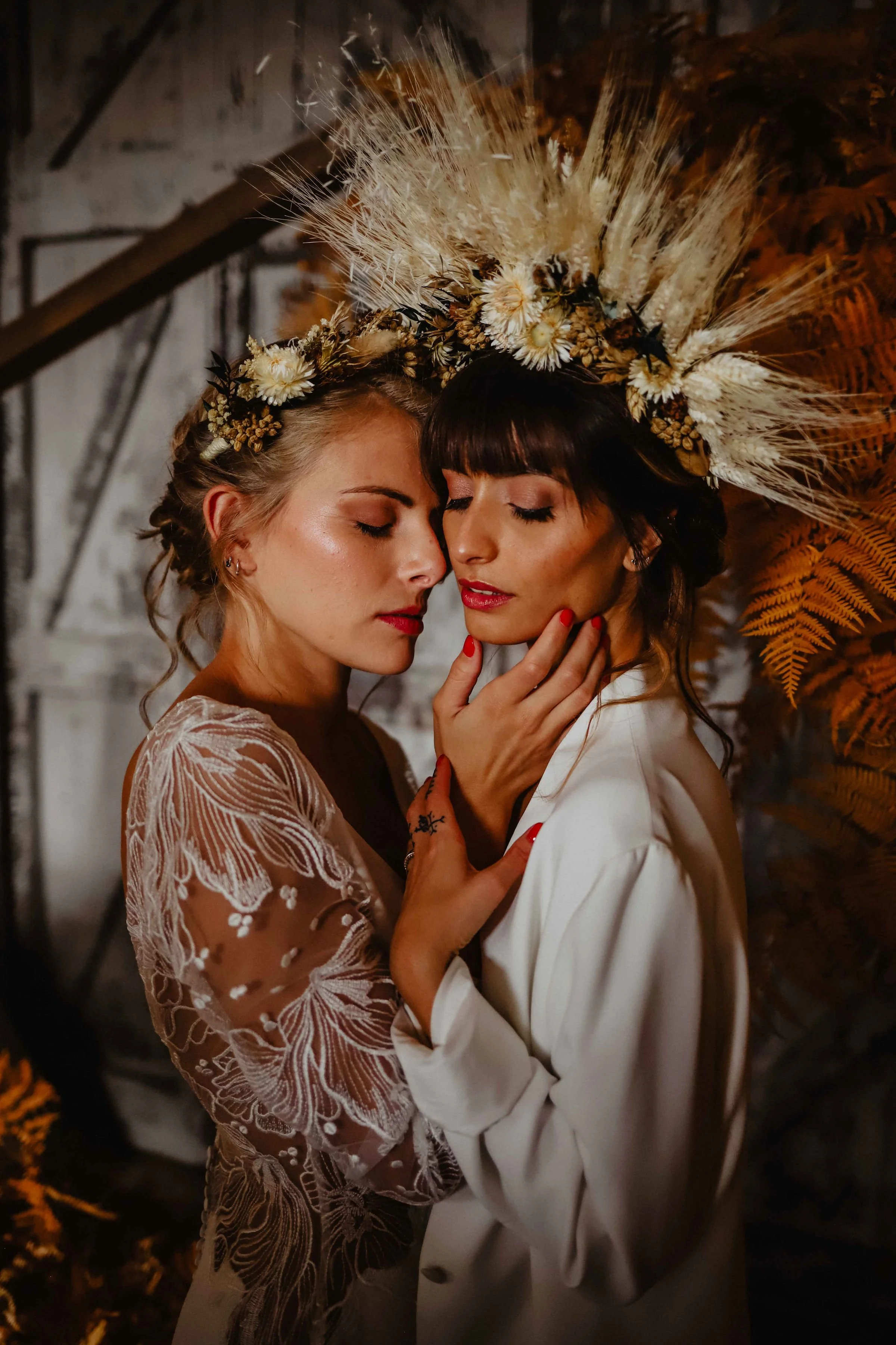 Deux femmes élégantes portent des couronnes de fleurs et se tiennent très près, avec les yeux fermés, dans une scène intime et chaleureuse.
