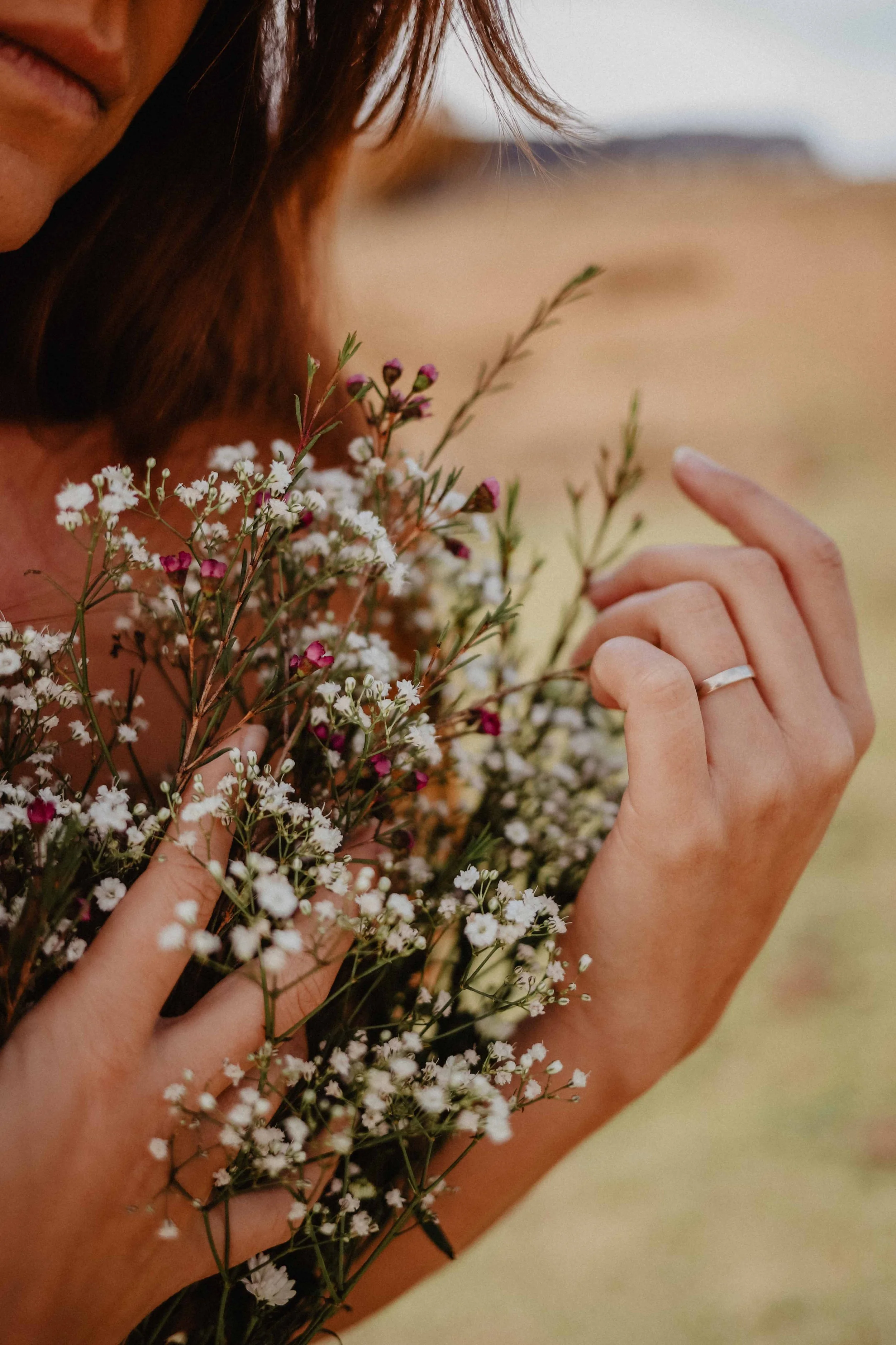 Une personne tient un bouquet de petites fleurs blanches et roses. On peut voir une main avec une alliance et un fond flou d'une zone naturelle.