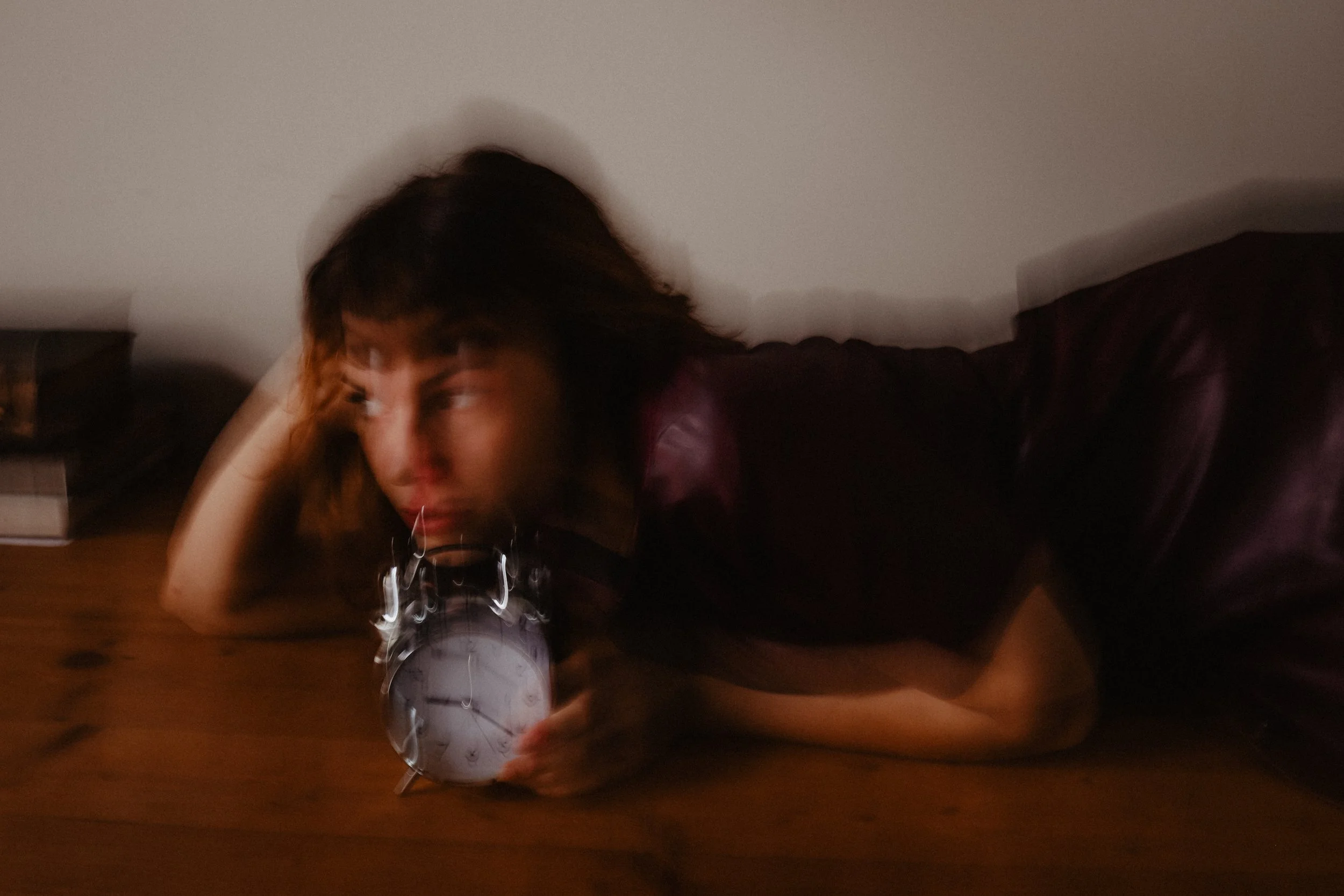 Une femme allongée sur une table en bois, tenant un réveil classique en métal dans sa main. La scène est floue, avec un éclairage tamisé et un fond neutre.