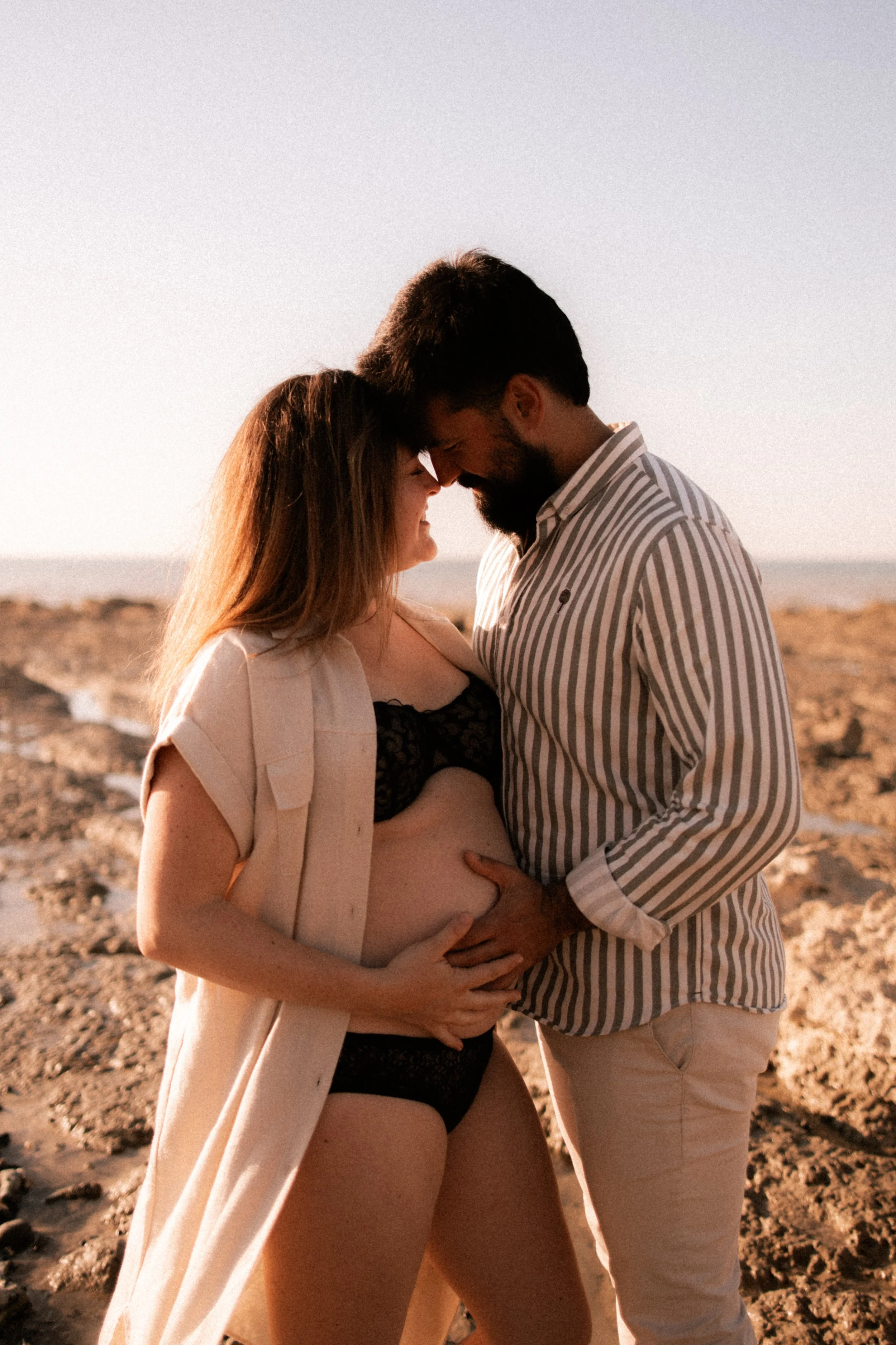 Un couple enceinte se tient sur une plage, se touchant le ventre et se dévisageant tendrement.