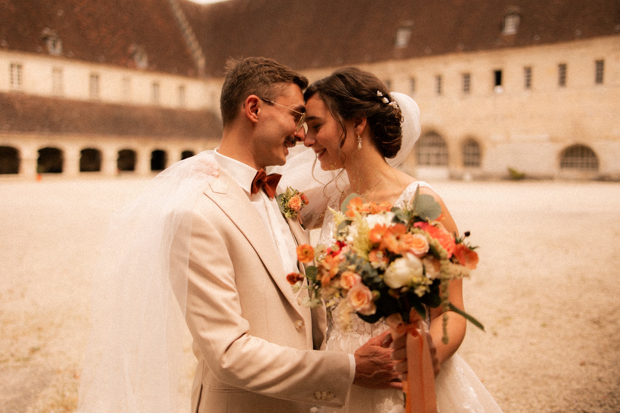 Un couple de mariés s'embrassant doucement devant un bâtiment ancien en pierre, la mariée tenant un bouquet de fleurs colorées.