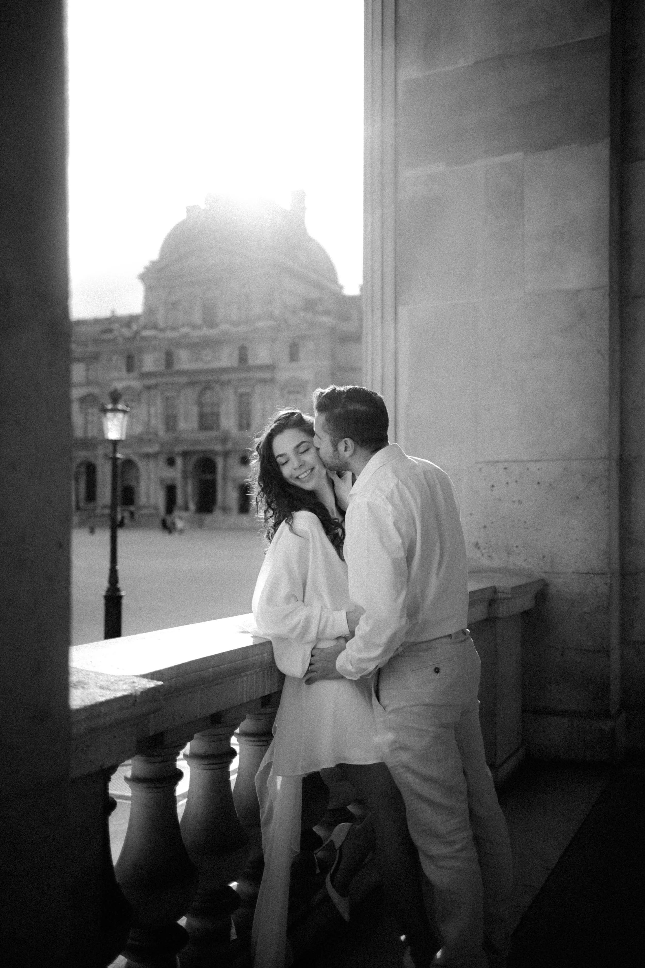 Un couple romantique s'embrassant sur une terrasse avec une vue sur un bâtiment historique devant le soleil couchant. Photographie en noir et blanc.