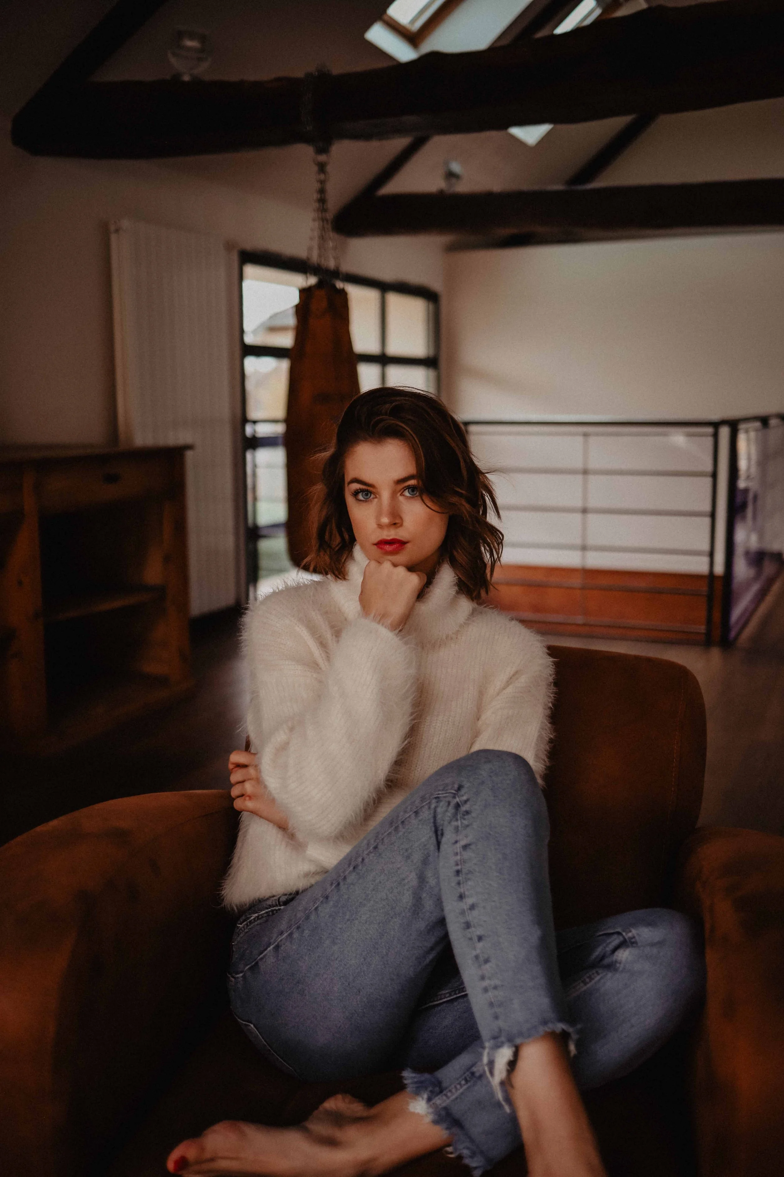 Une jeune femme assise dans un fauteuil en cuir, portant un pull blanc et des jeans déchirés, dans une pièce avec un intérieur en bois et un plafond en pente avec des poutres apparentes.