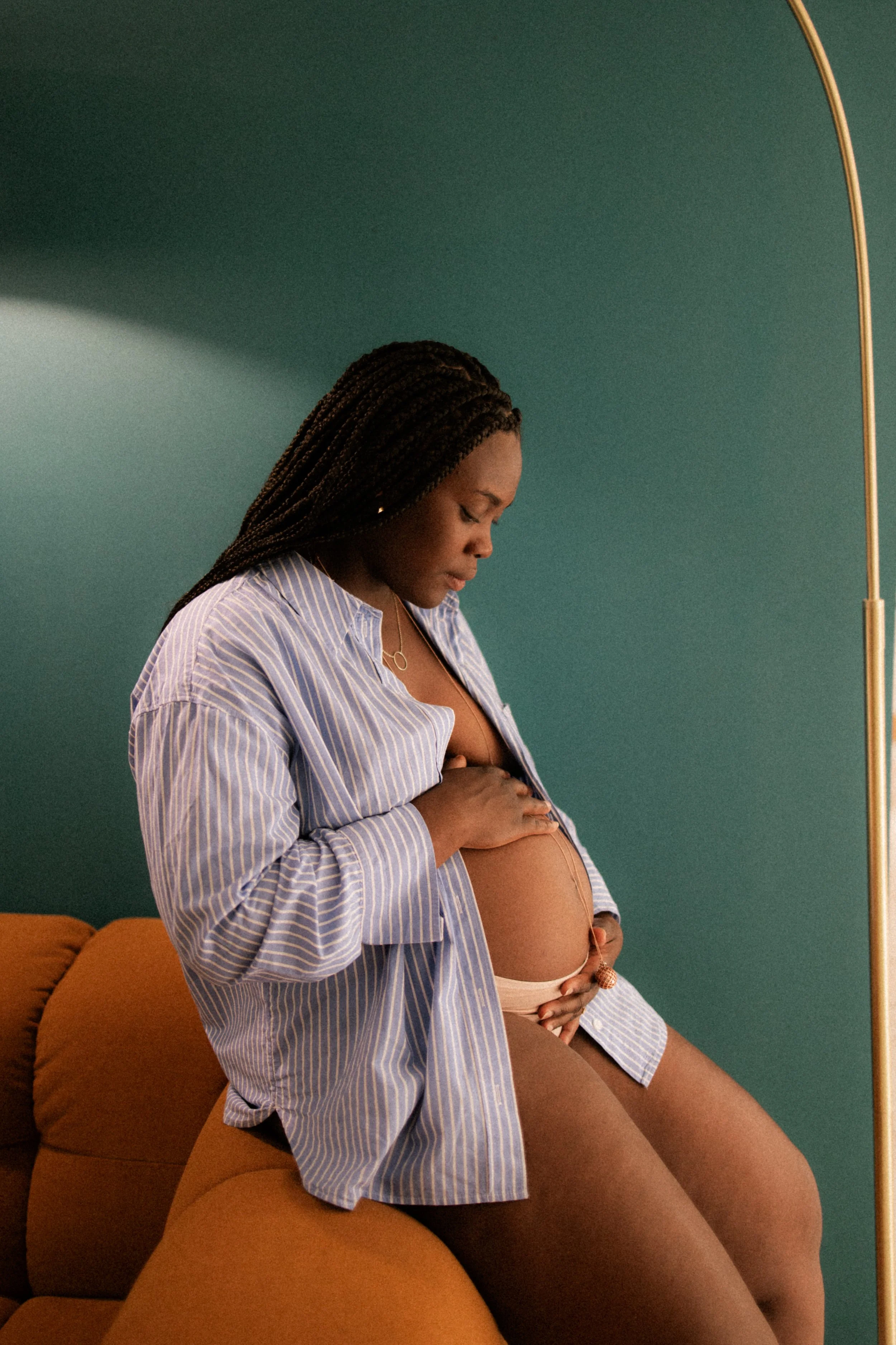 Femme enceinte assise sur un canapé orange, portant une chemise à rayures bleues et blanches, touchant son ventre avec une main. Mur vert en arrière-plan, lampe dorée à droite.
