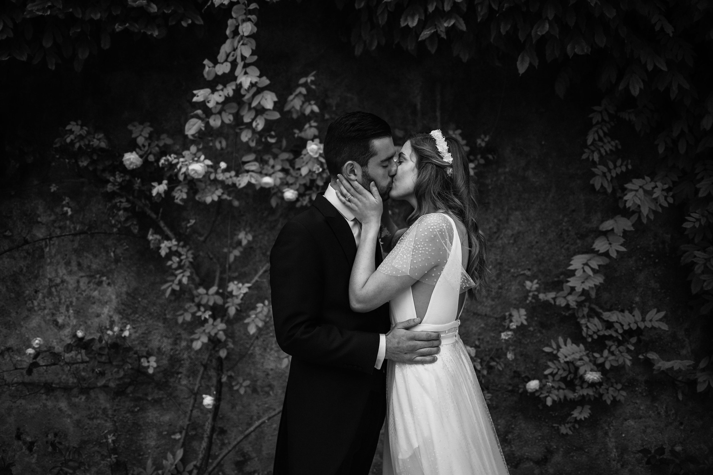 Un couple en mariage s'embrassant contre un mur orné de feuillage et de fleurs.