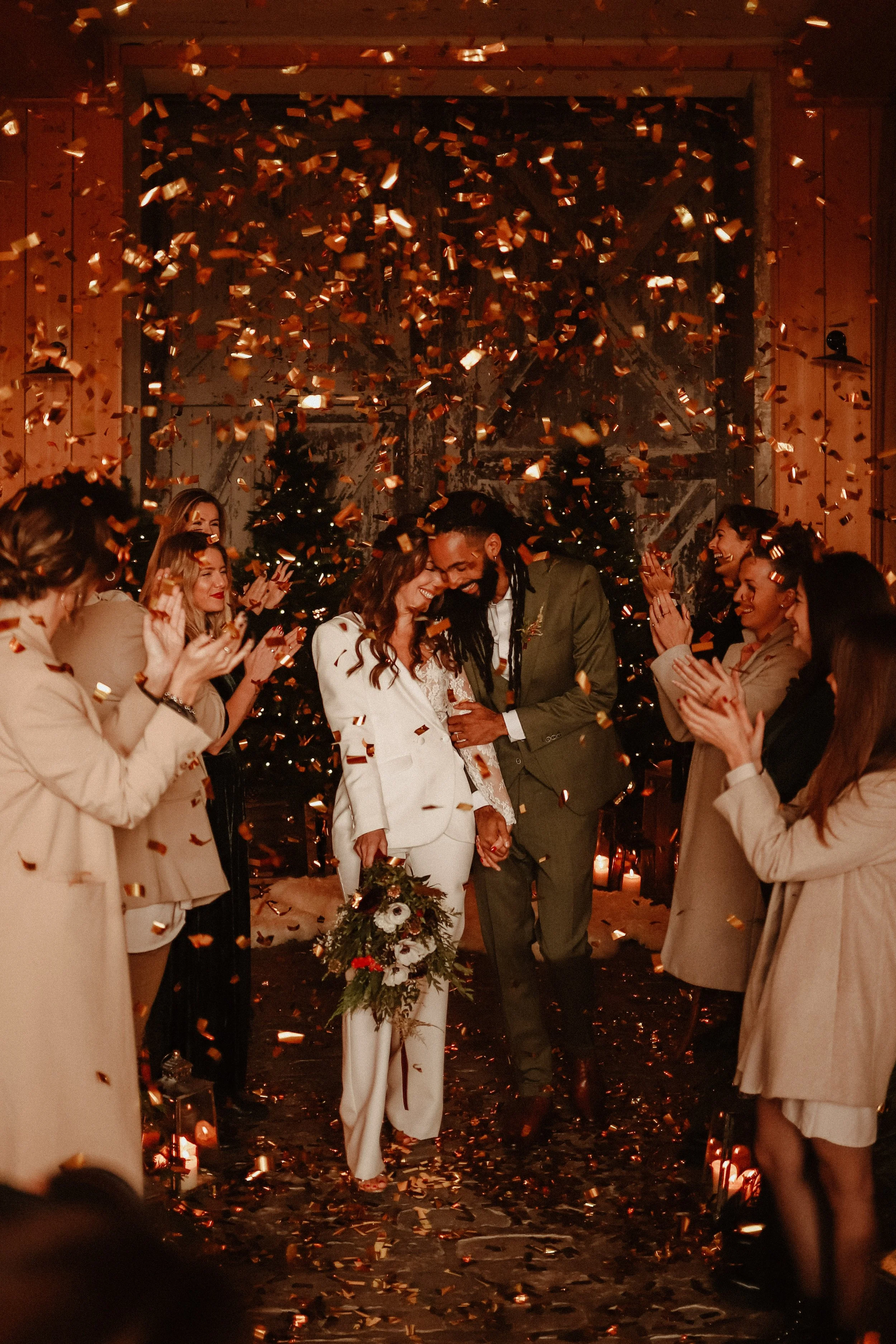 Un couple de mariés souriants, main dans la main, entourés d'invités applaudissant et lançant des confettis lors de leur mariage, avec des sapins de Noël en arrière-plan.
