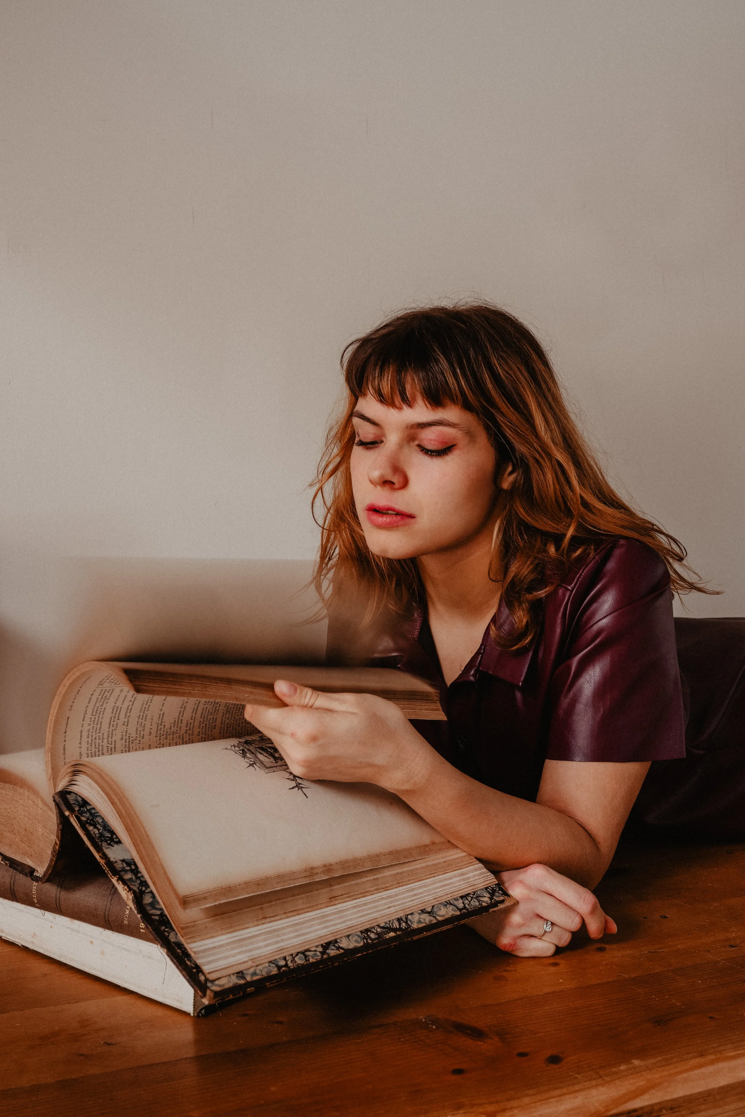 Une femme aux cheveux roux, portant une veste en cuir violette, lisant un grand livre posé sur une table en bois.