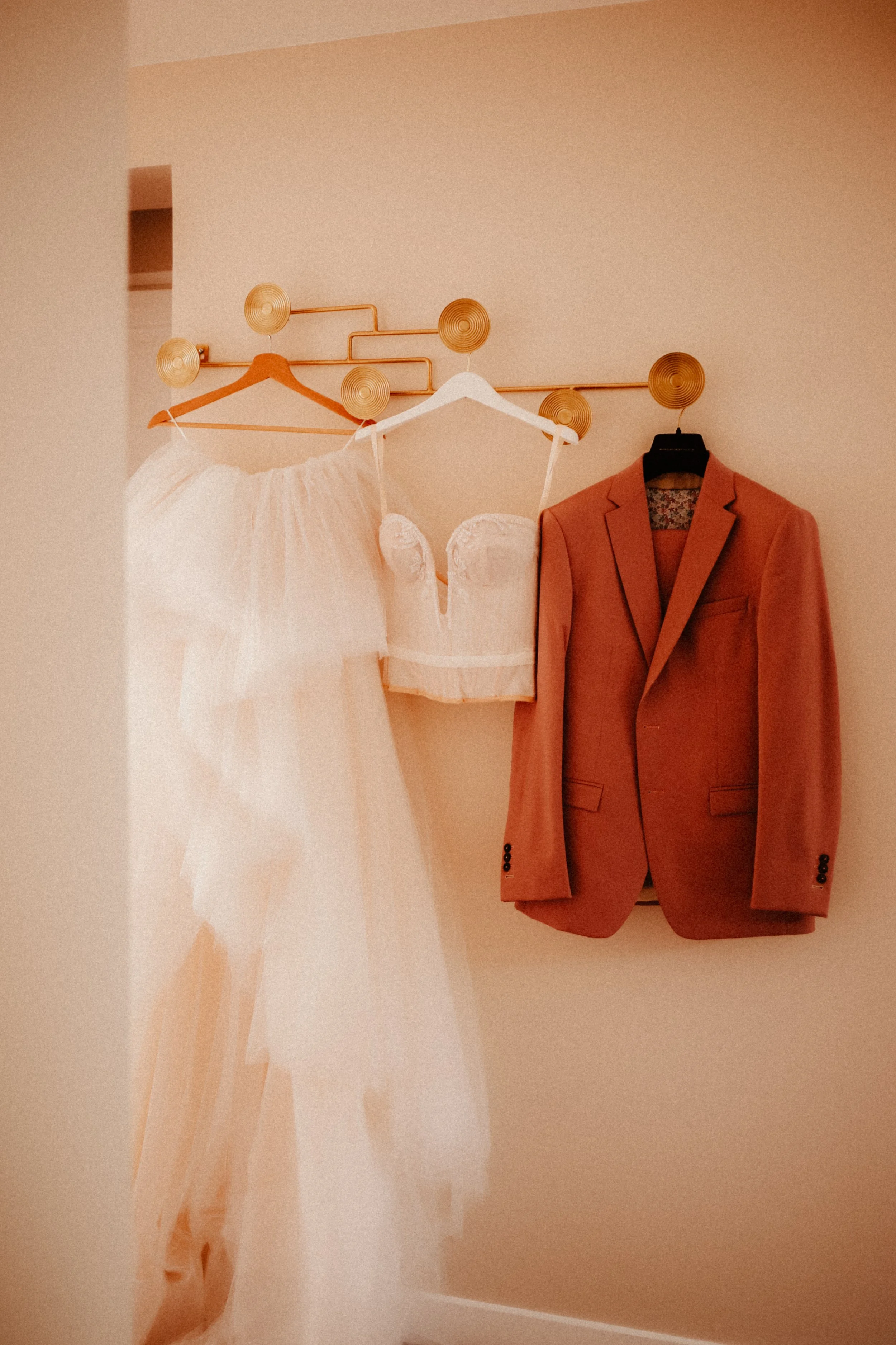 Robe de mariée blanche, bustier en velours blanc, veste de costume rose sur un mur beige.
