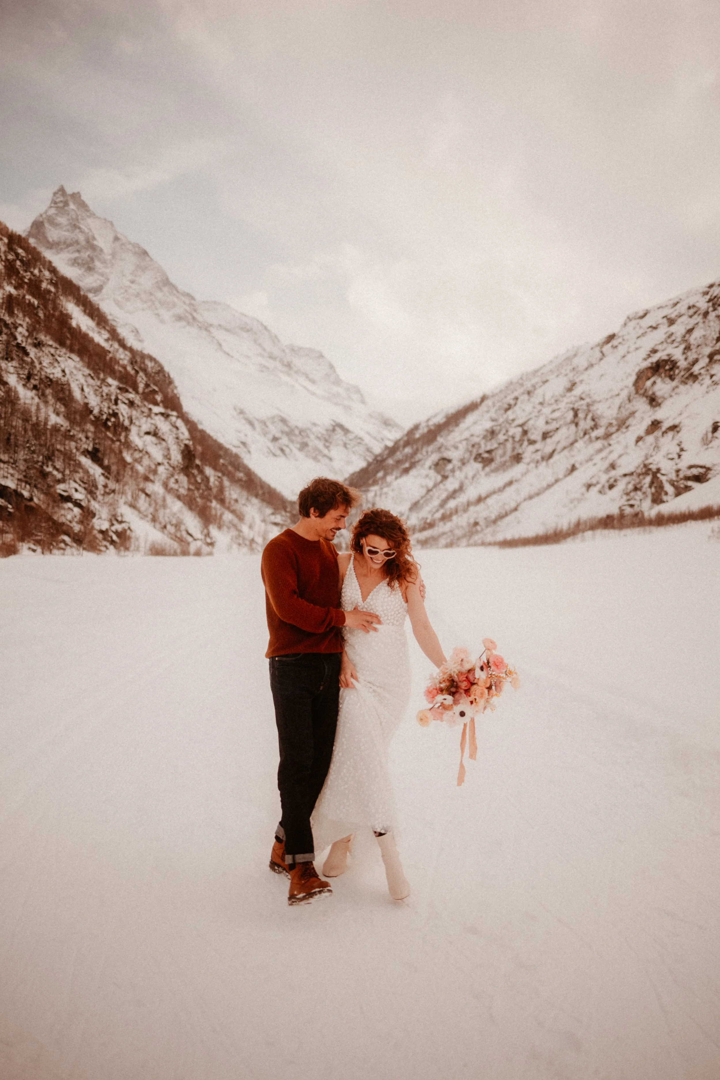 Un couple en tenue de mariage se promène dans la neige avec des montagnes en arrière-plan, la femme porte une robe blanche et des lunettes de soleil, tenant un bouquet de fleurs.