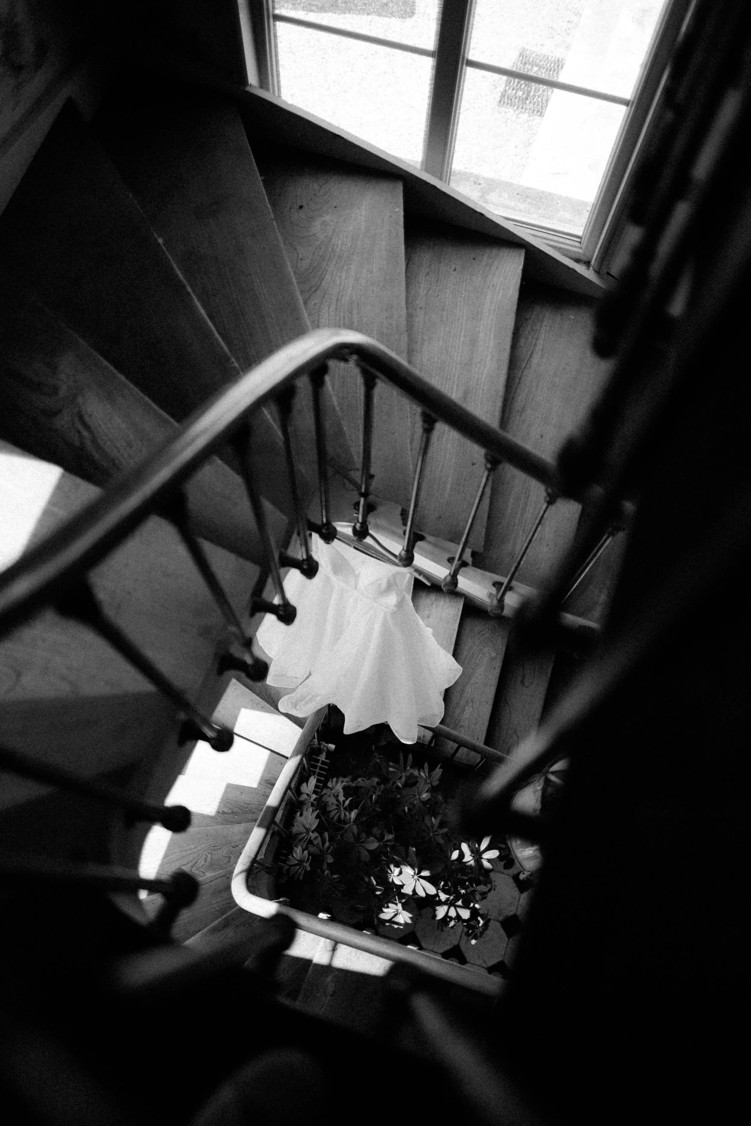 Vue du haut d'un escalier en bois avec une rampe en métal, un rideau blanc au bas de la cage d'escalier et une fenêtre laissant entrer la lumière.