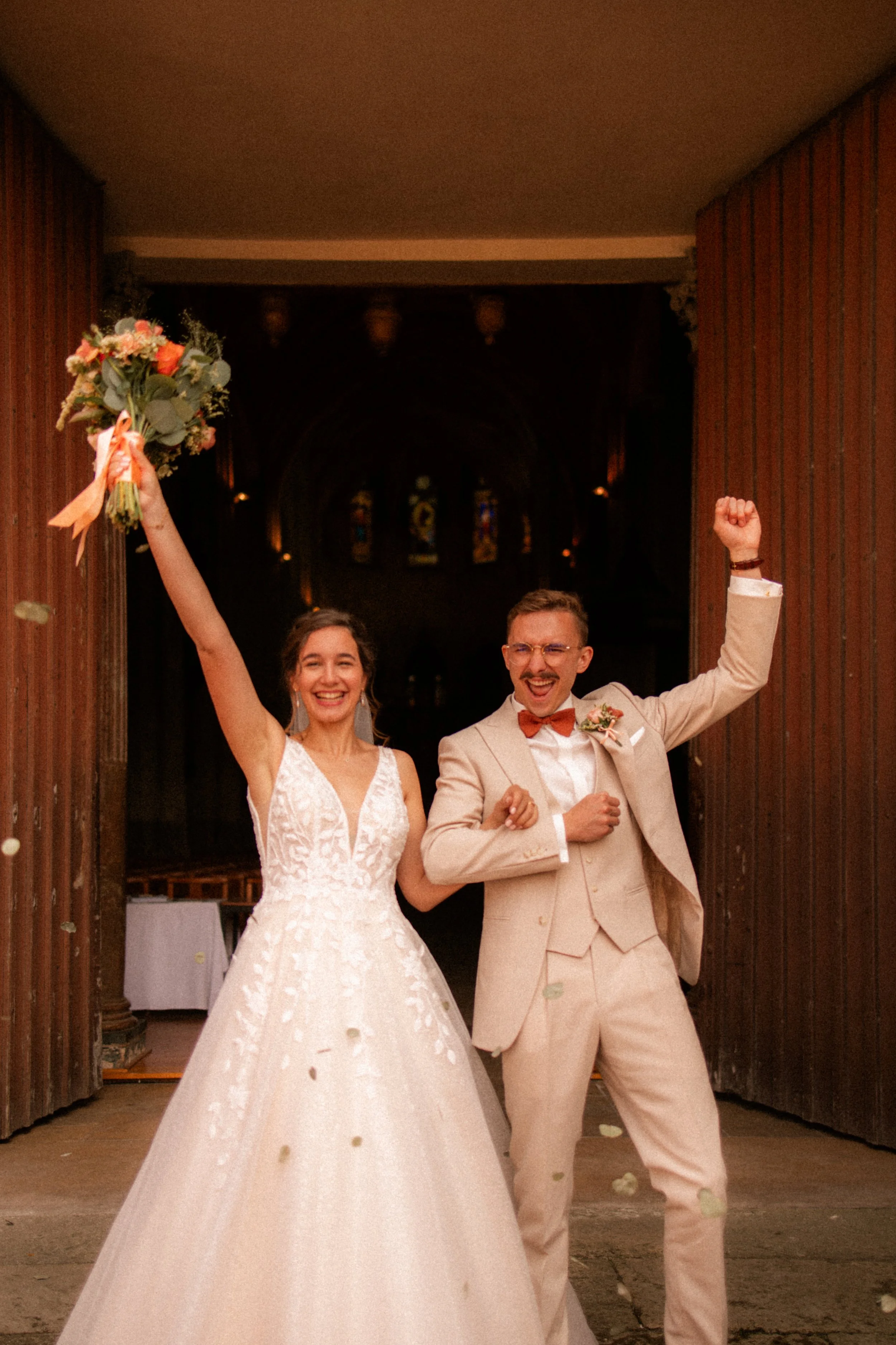 Jeunes mariés heureux, célébrant leur mariage, avec la mariée tenant un bouquet de fleurs en levant le bras et le marié faisant un geste de victoire avec le poing.