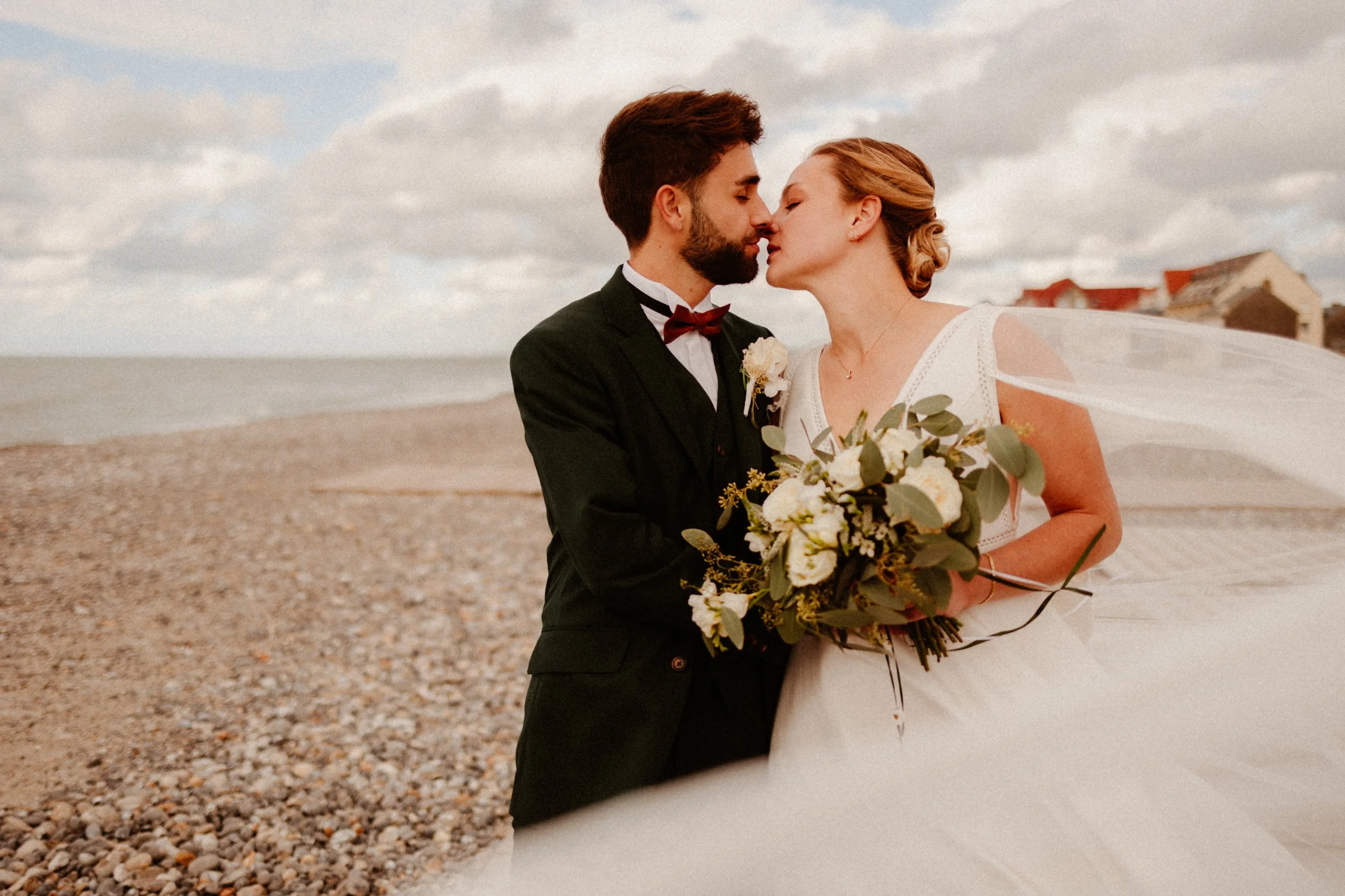 Un couple de mariés se tenant près de la mer, se préparant à s'embrasser, le brouillon et les nuages dans le ciel en arrière-plan.