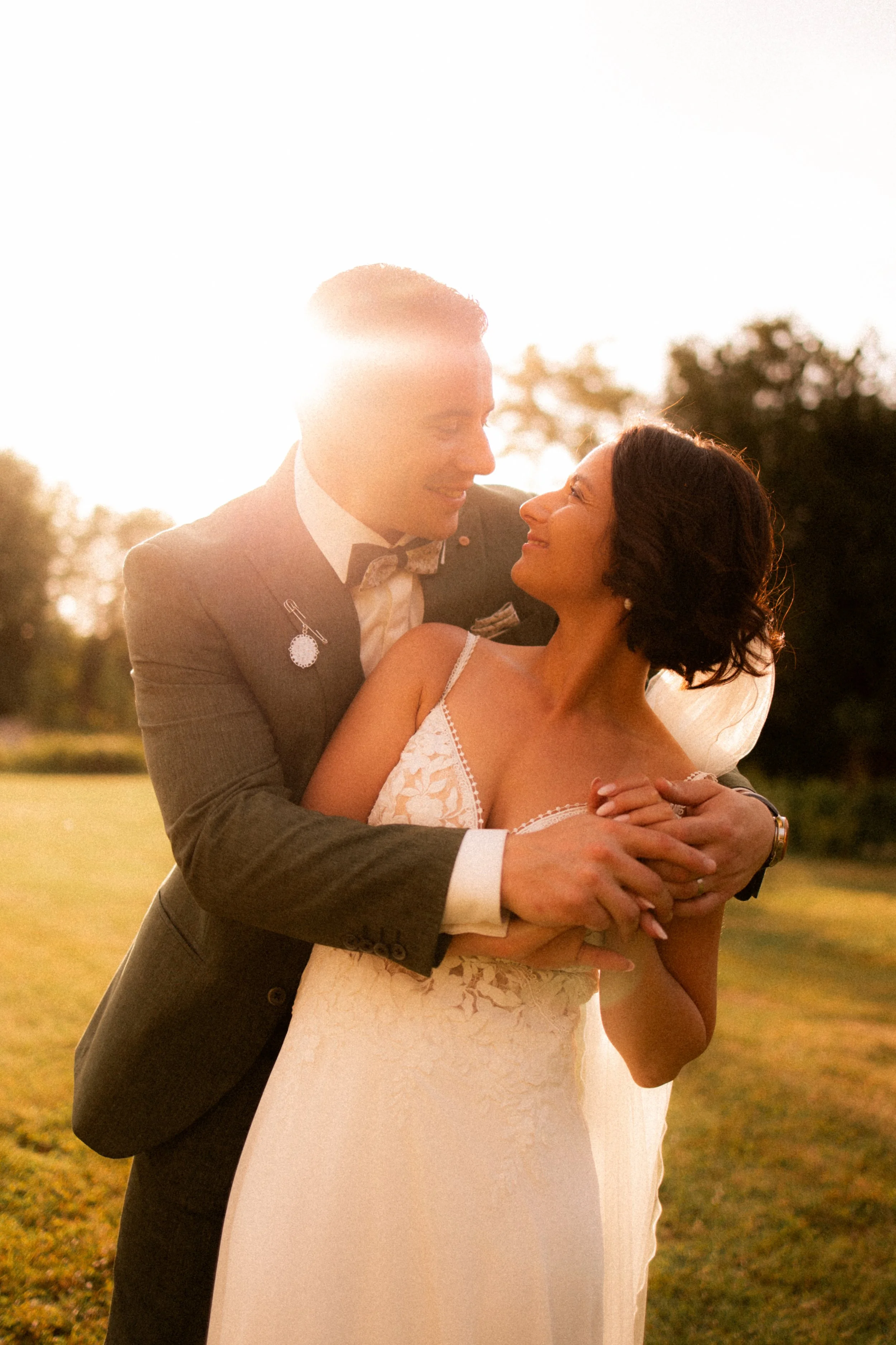 Un couple de mariés s'enlace dans un champ lors d'un coucher de soleil, le marié en costume et la mariée en robe de mariage, ambiance romantique.