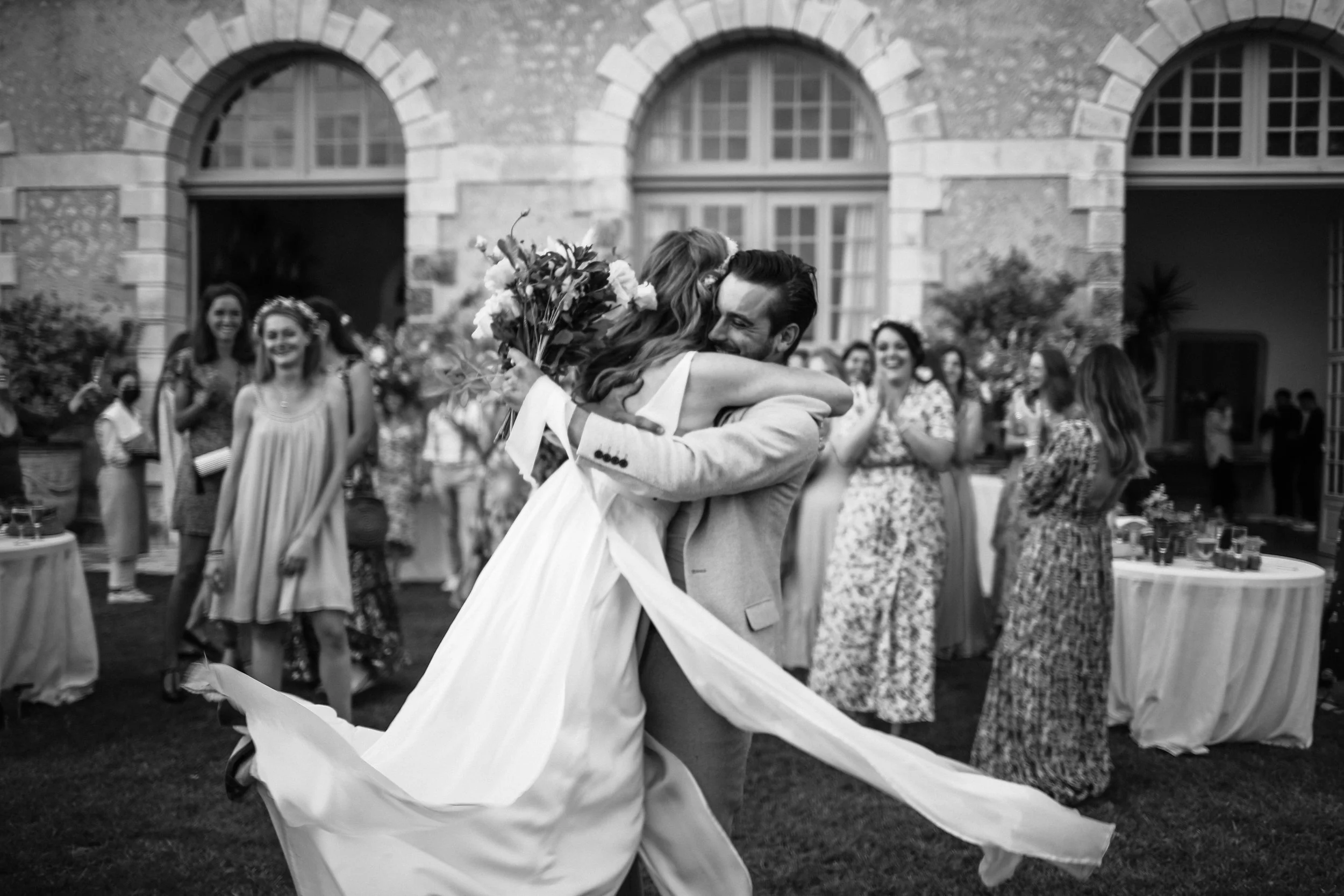 Un couple d'amis qui se retrouve pour une embrassade dans un cadre de mariage, avec des invités souriants en arrière-plan, dans une ambiance festive en extérieur.