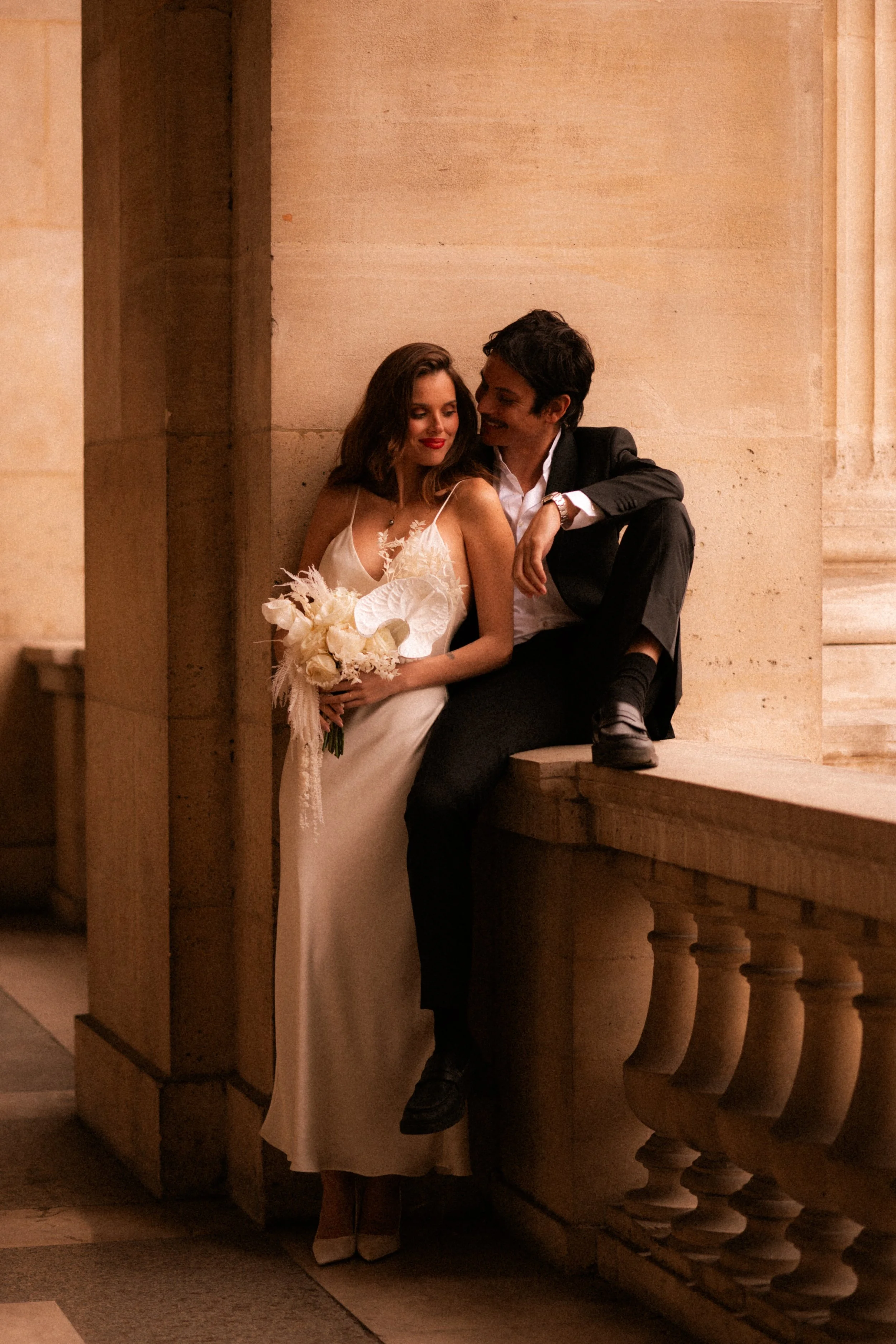 Un couple habillé élégamment, la femme en robe de mariage blanche et l'homme en costume noir, assis contre une paroi en pierre dans un lieu architectural ancien, semblant se détendre et se regarder avec affection.