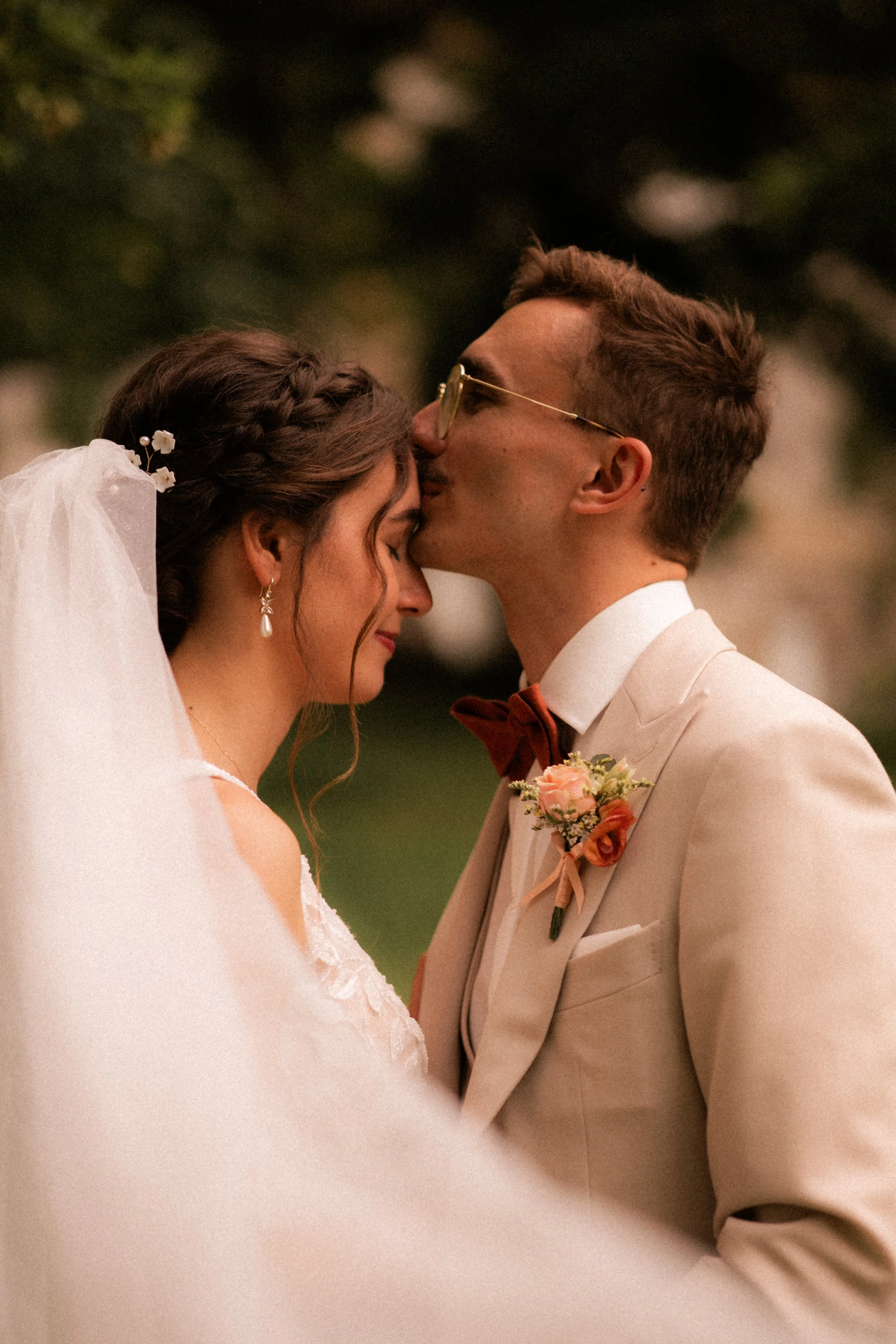 Un couple de mariés lors d'une cérémonie, avec la femme portant une robe blanche avec voilage, des boucles d'oreilles en perles, et une coiffure tressée décorée de fleurs, et l'homme en kosten beige, chemise blanche et nœud papillon rouge, pendant qu