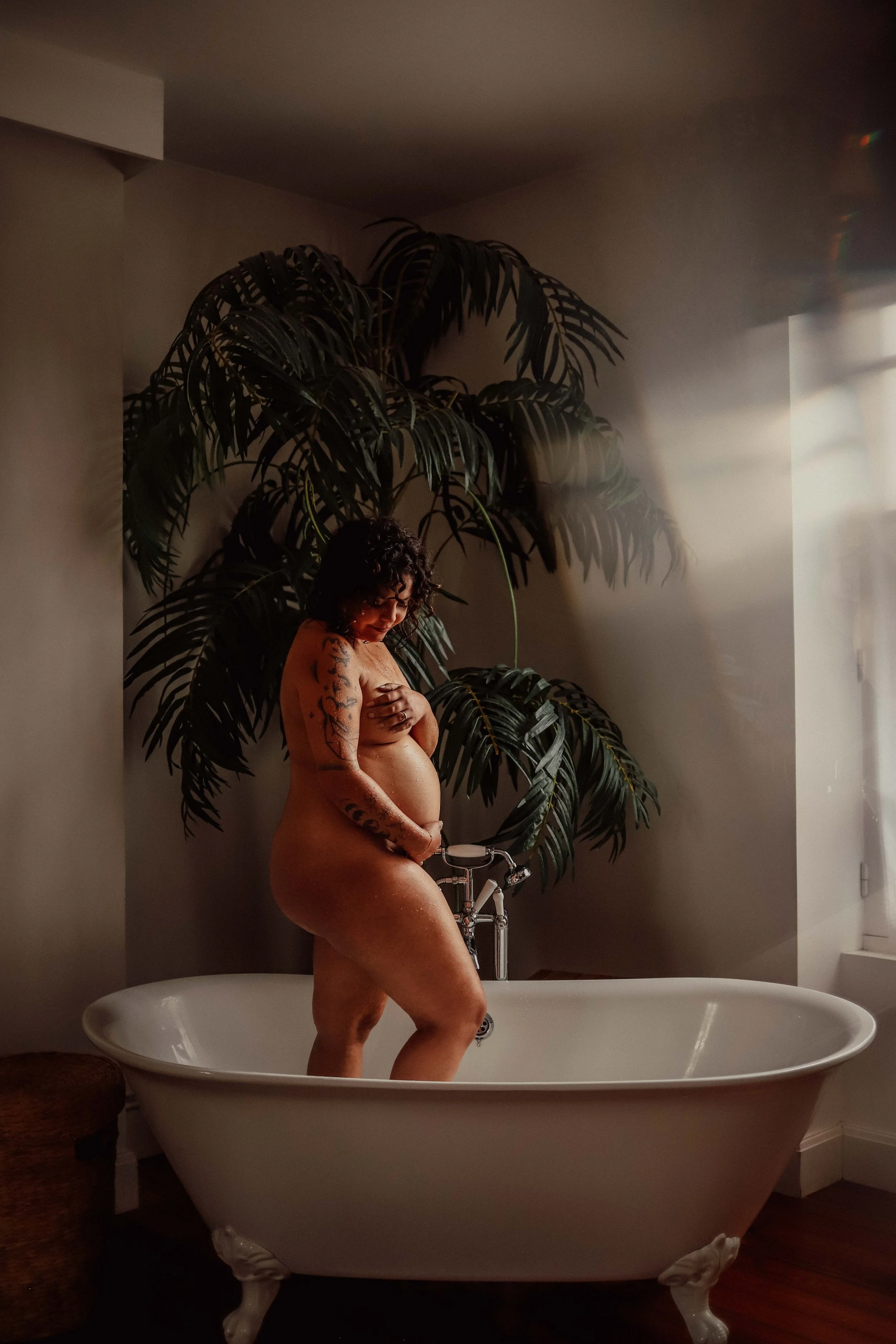 Une femme nue avec des tatouages se tient dans une baignoire en pleine lumière naturelle, avec un grand plant derrière elle dans une pièce lumineuse.