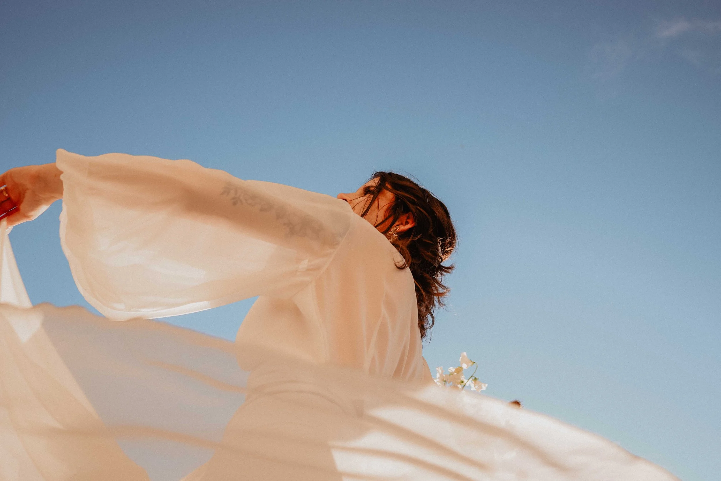 Une femme vêtue d'une robe légère blanche ou crème, levant les bras, sous un ciel bleu clair.