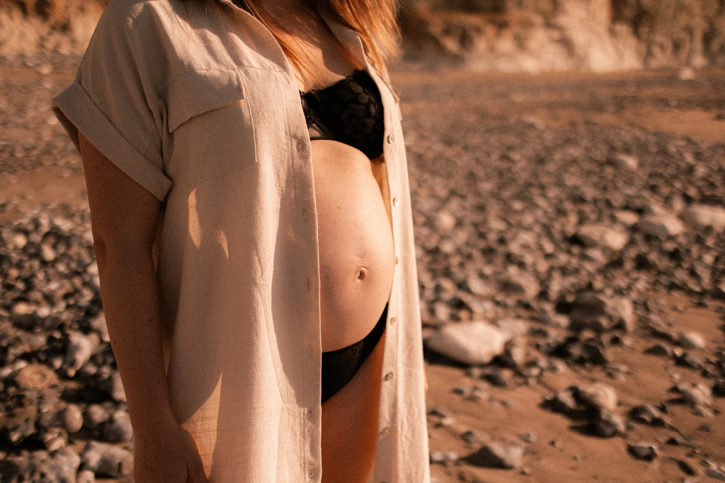 Une femme enceinte portant un haut noir et un long manteau beige examine son ventre à l'extérieur, sur une plage rocheuse pendant le coucher de soleil.