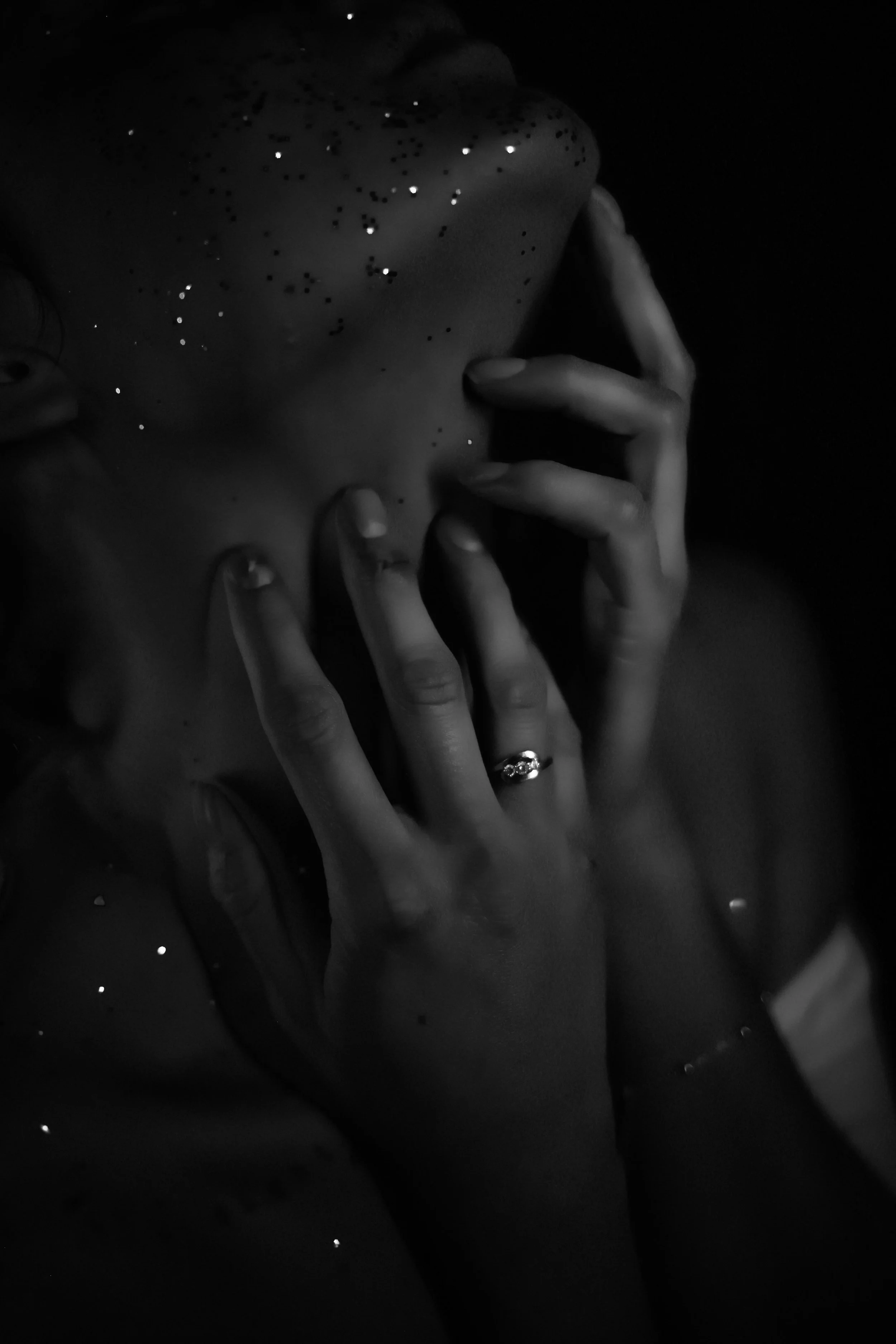 Une personne en noir et blanc, avec des mains près du visage, portant une bague, et des paillettes sur la peau, dans un décor sombre.