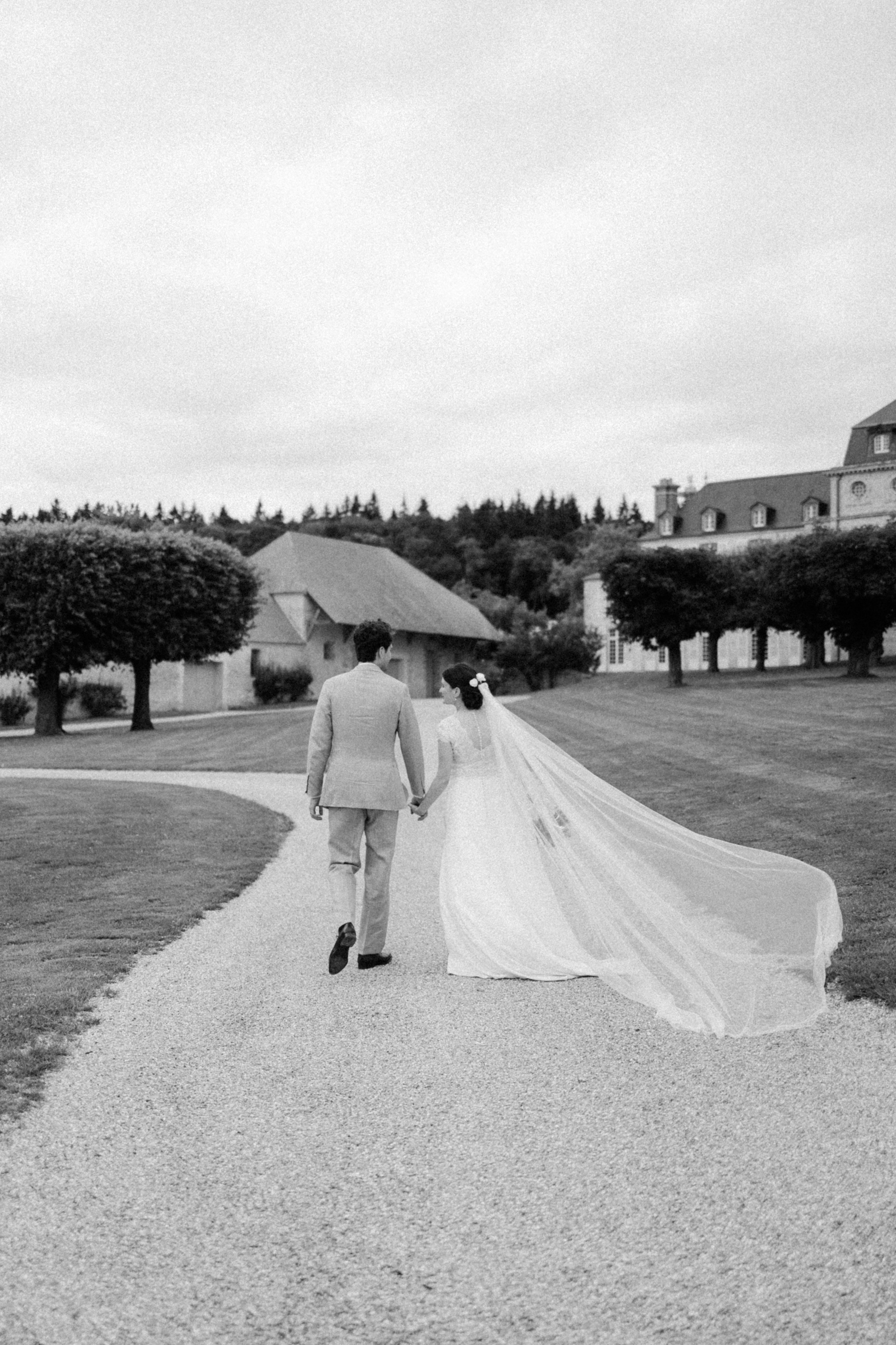 Un couple de mariés marche main dans la main sur un chemin de gravier dans un paysage rural avec des arbres et des bâtiments en arrière-plan, en noir et blanc.